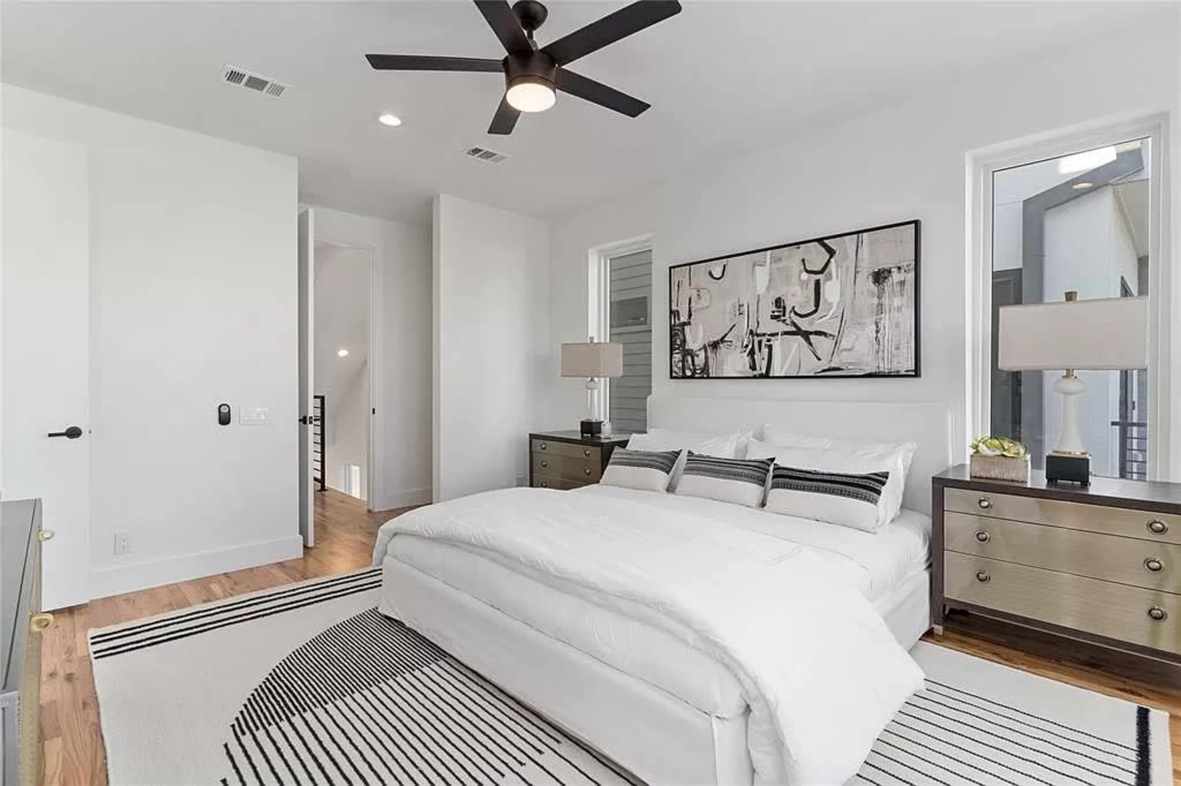 A modern bedroom features a white bed with a striped blanket, two bedside lamps, and abstract art on the walls.