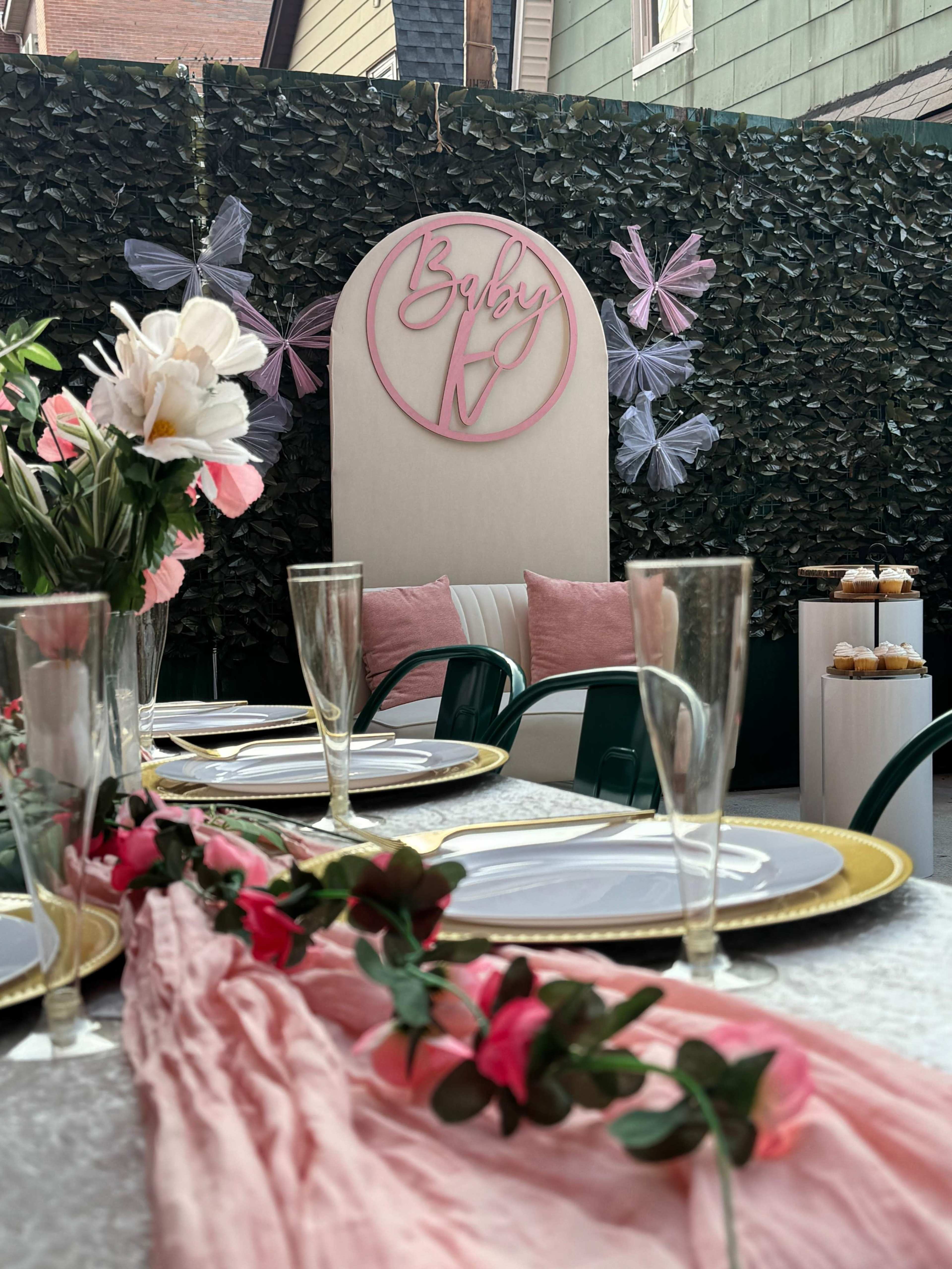 The image shows a decorated outdoor space with a table set for a celebration, featuring a floral arrangement, elegant tableware, and a backdrop with a sign that reads "Baby V."