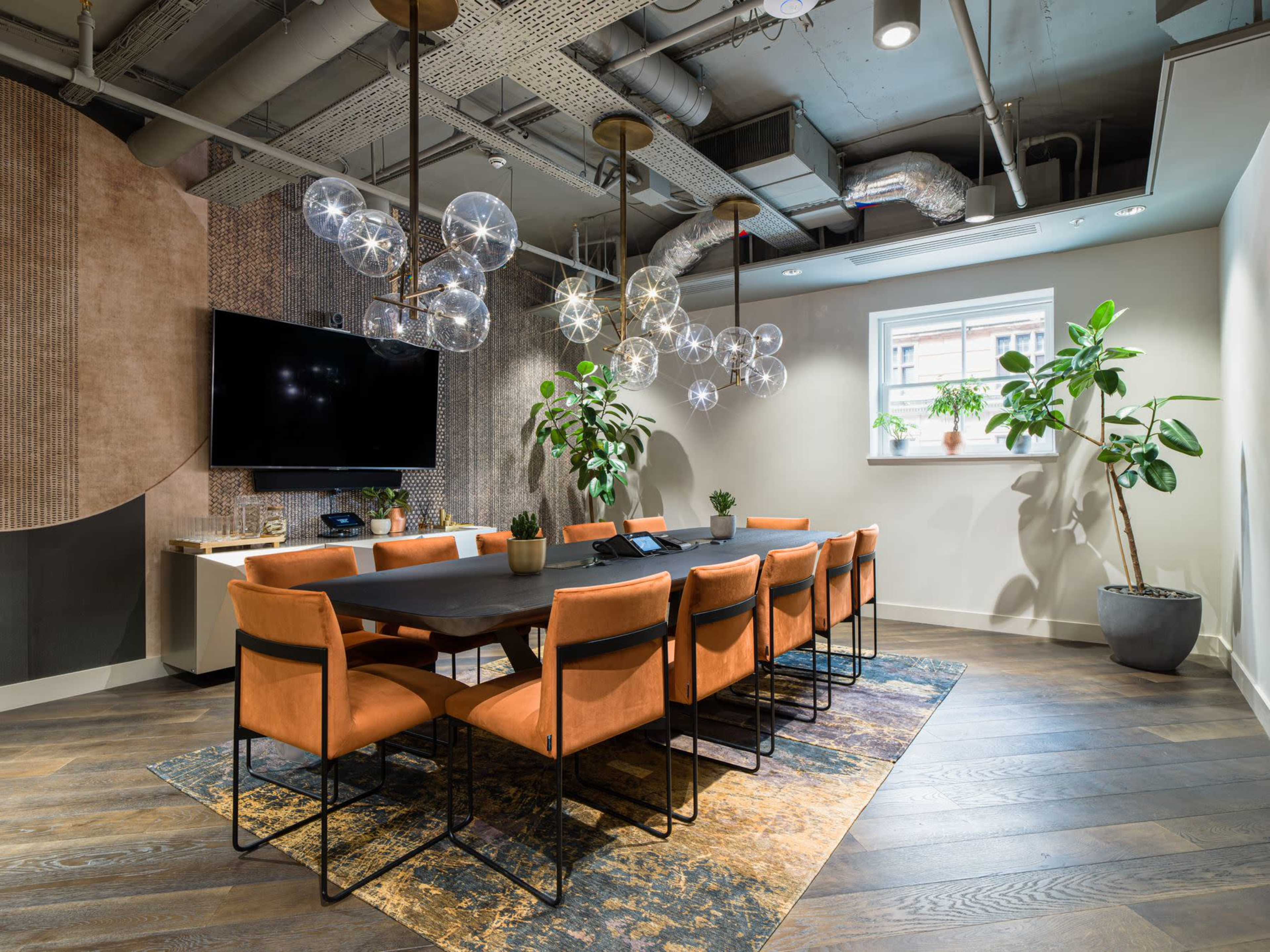 The image shows a modern conference room featuring a long black table surrounded by orange chairs, decorative plants, and a large screen mounted on the wall.
