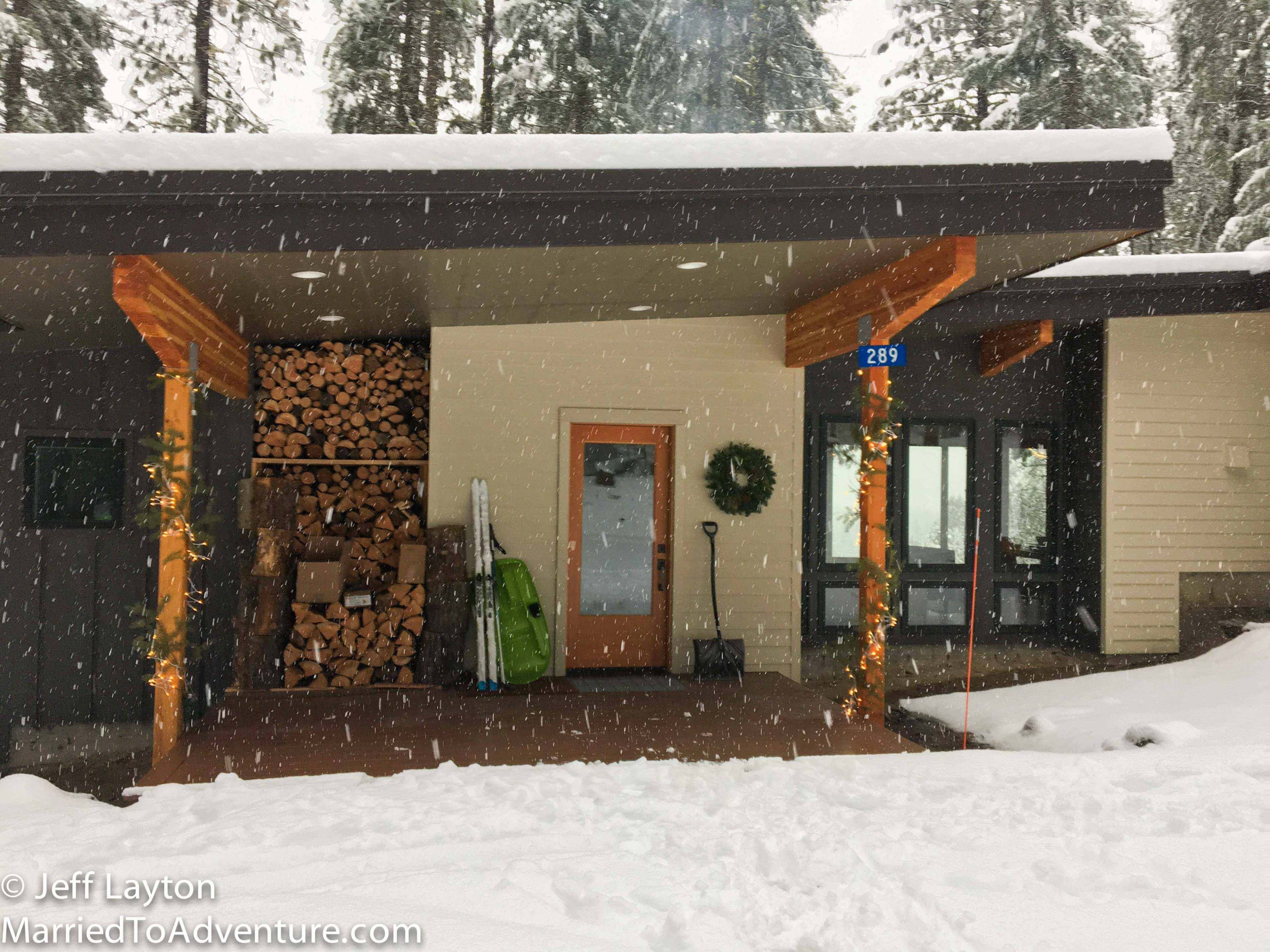 A modern house is situated in a snowy landscape, featuring a stacked woodpile and decorations along the porch.