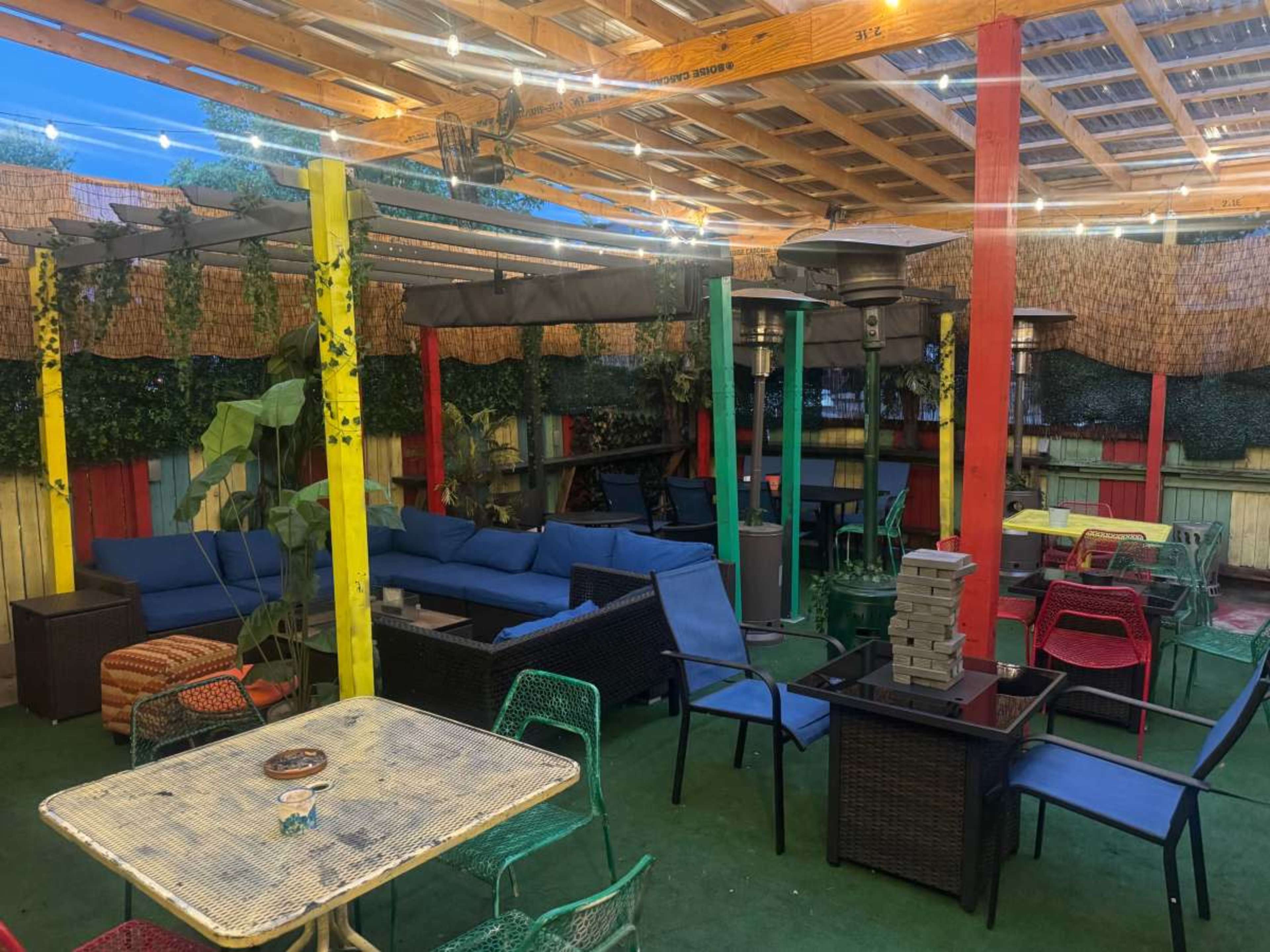 The image shows a colorful outdoor seating area with various types of furniture, including couches, tables, and heaters, under a wooden roof.