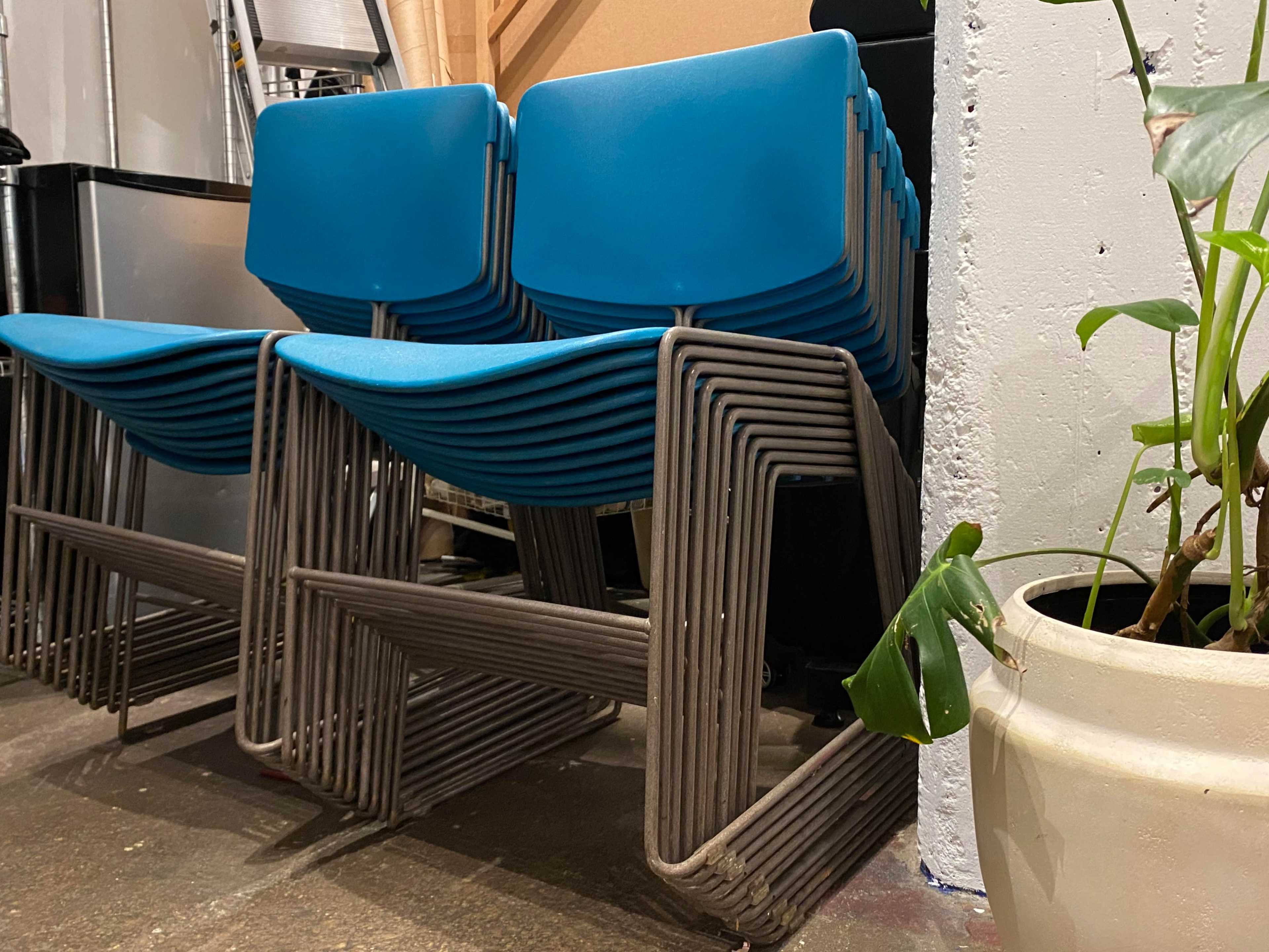 A stack of blue chairs is leaning against a wall next to a plant in a white pot.