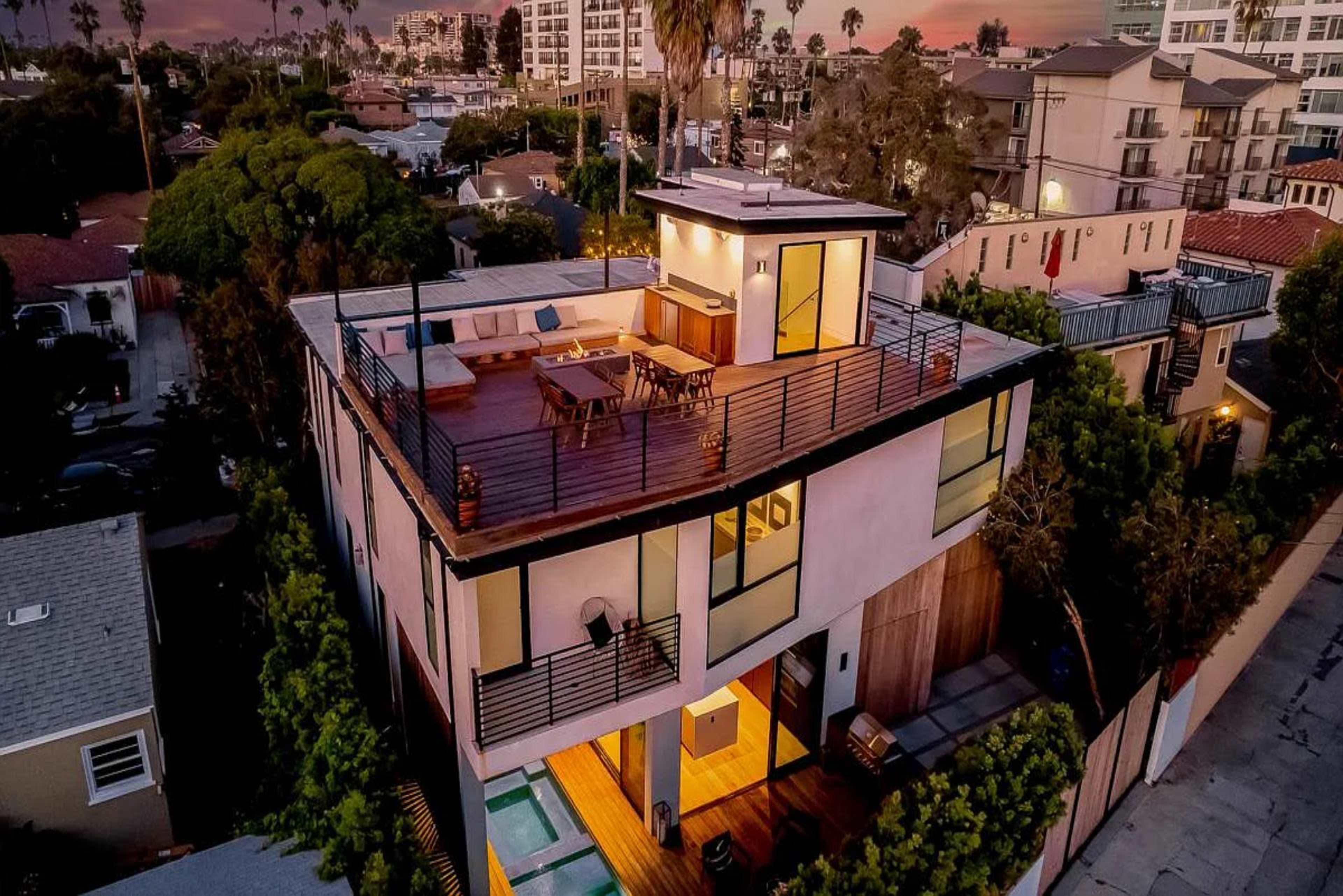 The image shows a modern two-story house with a rooftop deck, surrounded by trees and adjacent buildings in an urban setting.