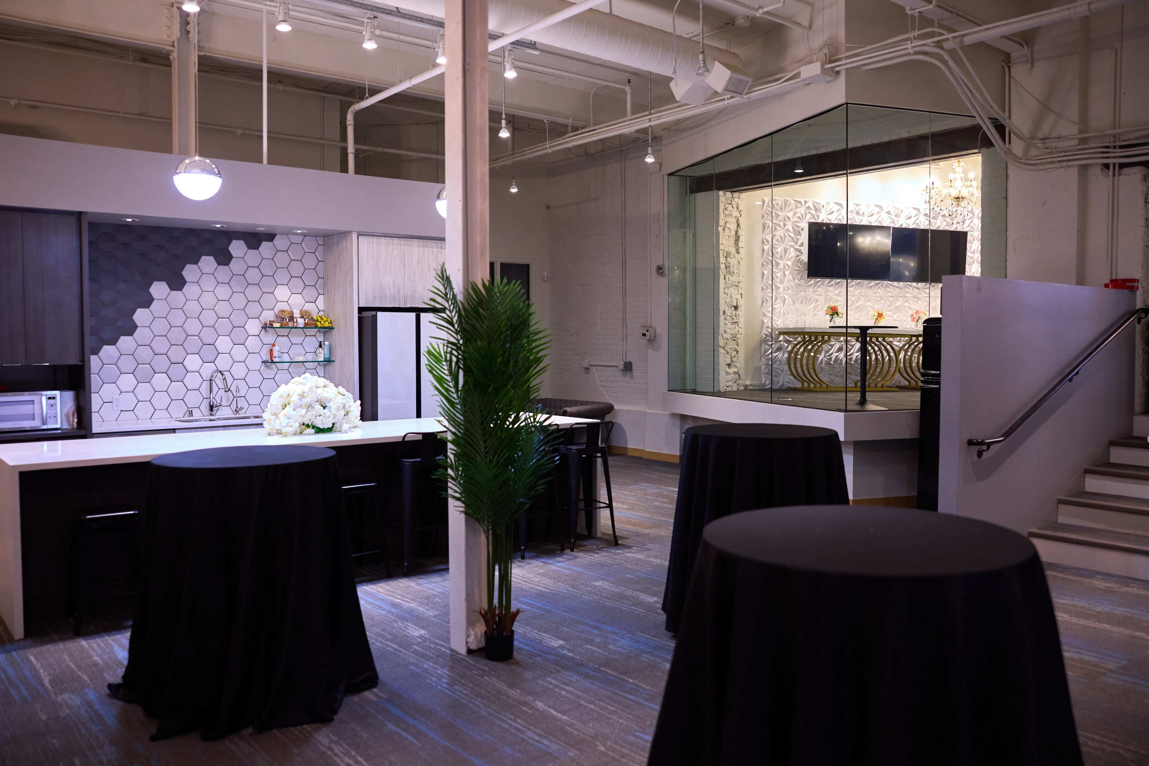 The image shows a modern kitchen and lounge area with high tables, a floral arrangement on the counter, and a glass-walled room in the background.