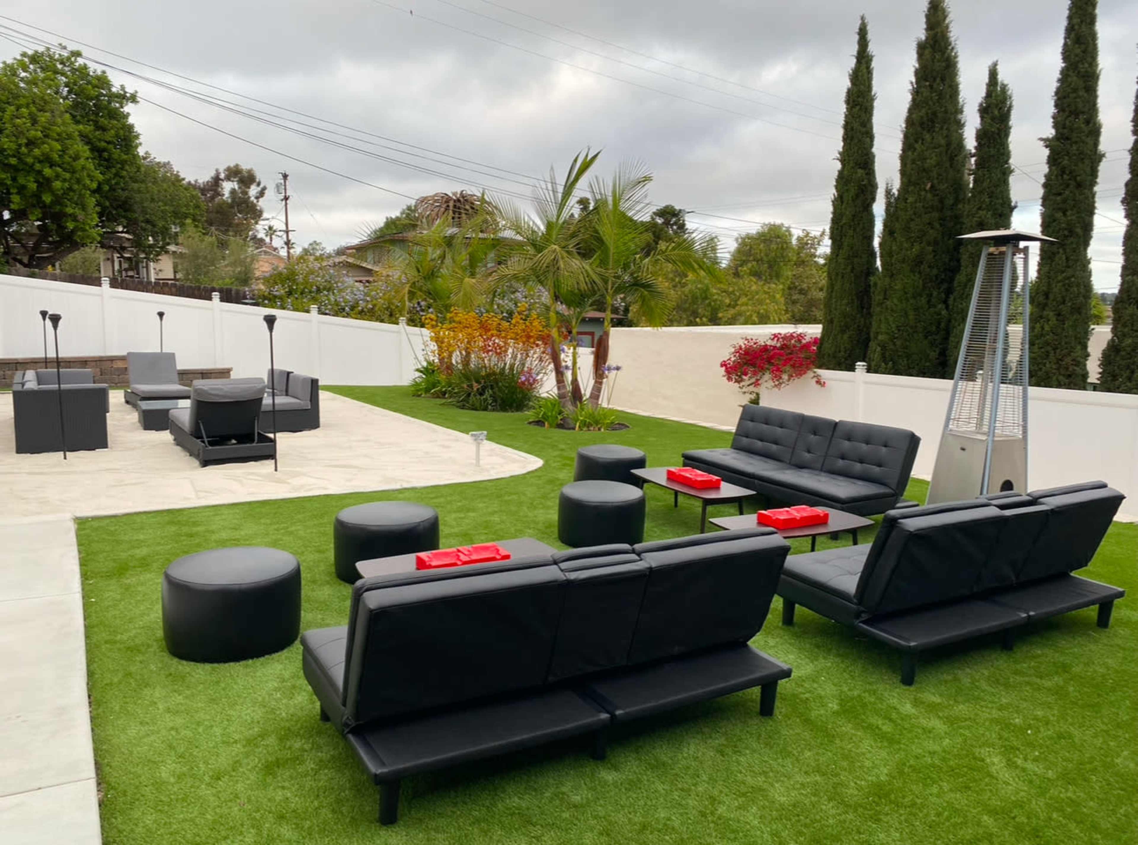 The image shows a modern outdoor seating area on green grass, featuring black couches, round tables, and a stone patio with potted plants and tall trees in the background.