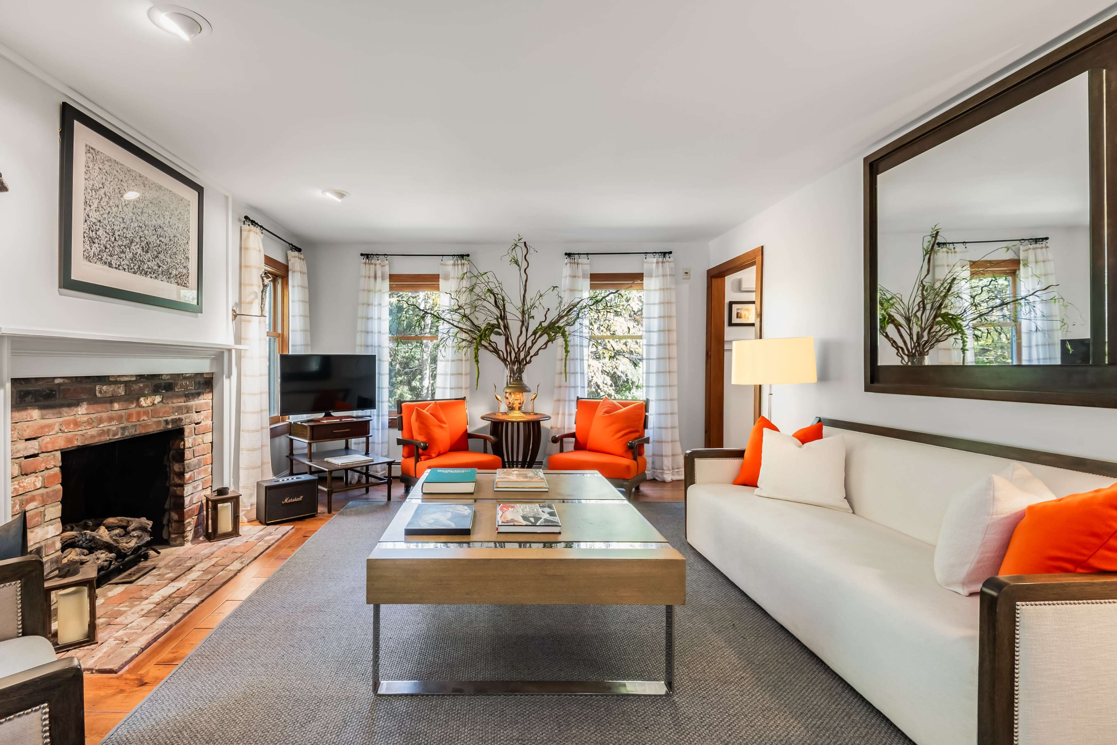 The living room features a white sofa and two orange chairs arranged around a metallic coffee table, with a brick fireplace and a large mirror on the wall.