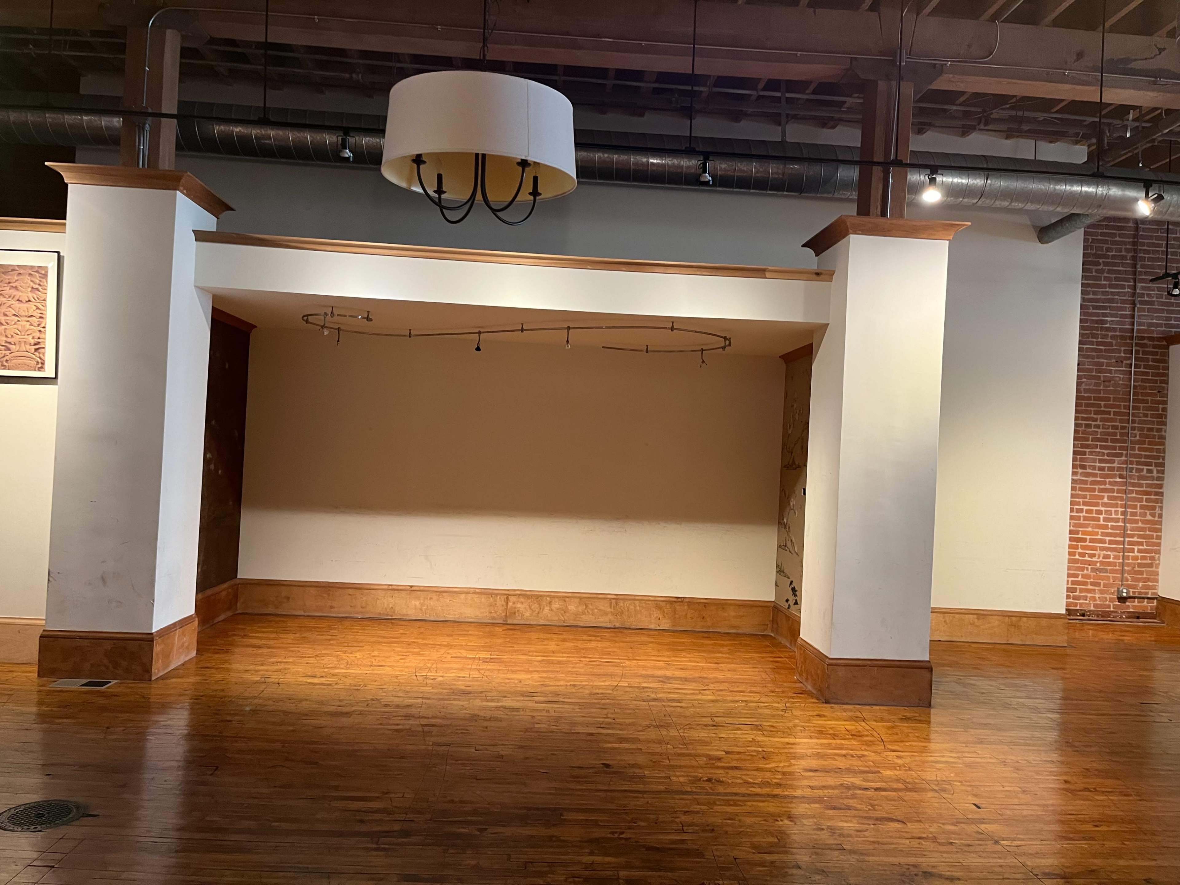 The image shows an empty interior space with wooden flooring, white walls, and a central hanging light fixture.