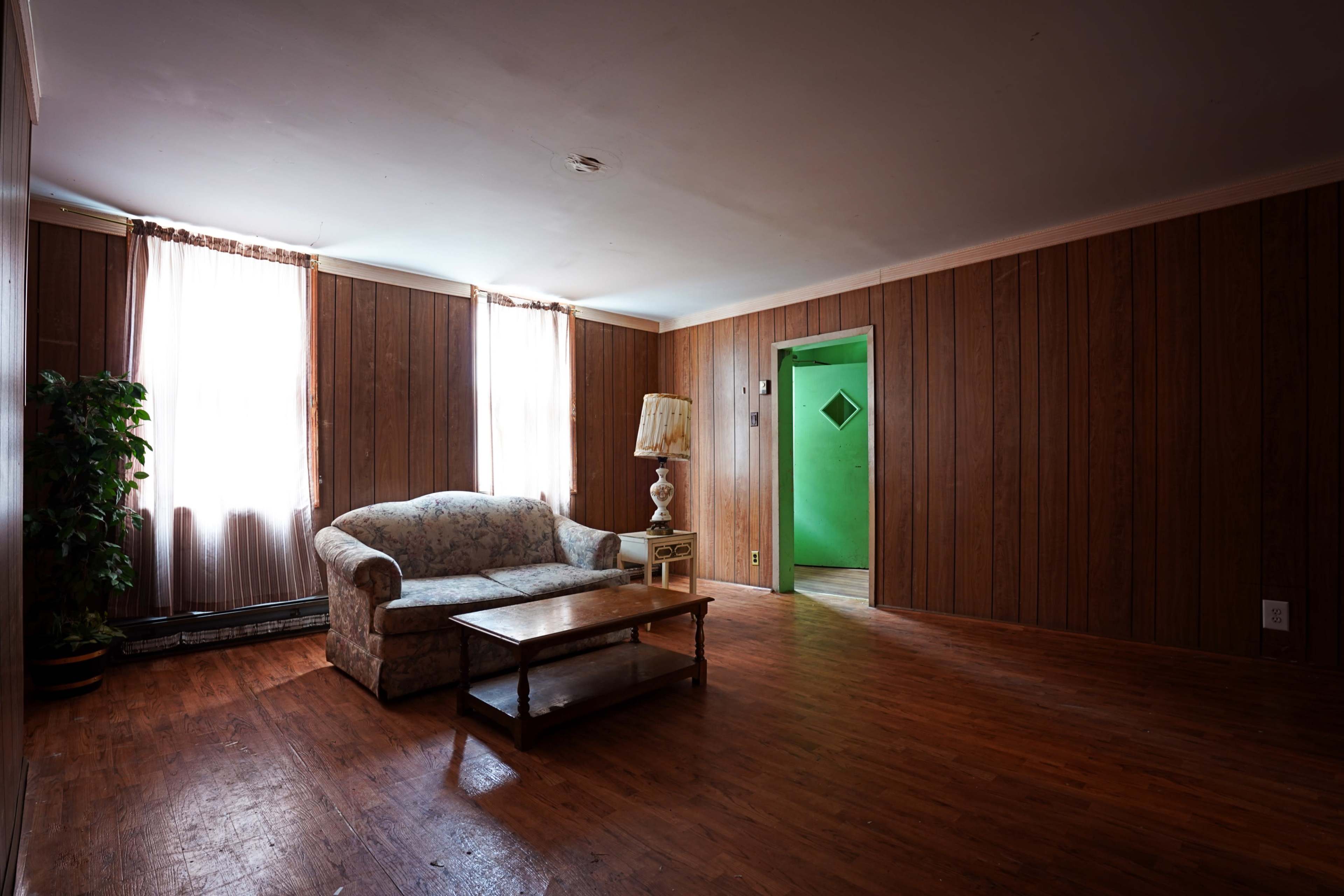 An empty living room with wood-paneled walls, a floral couch, a coffee table, and a green door leading to another room.