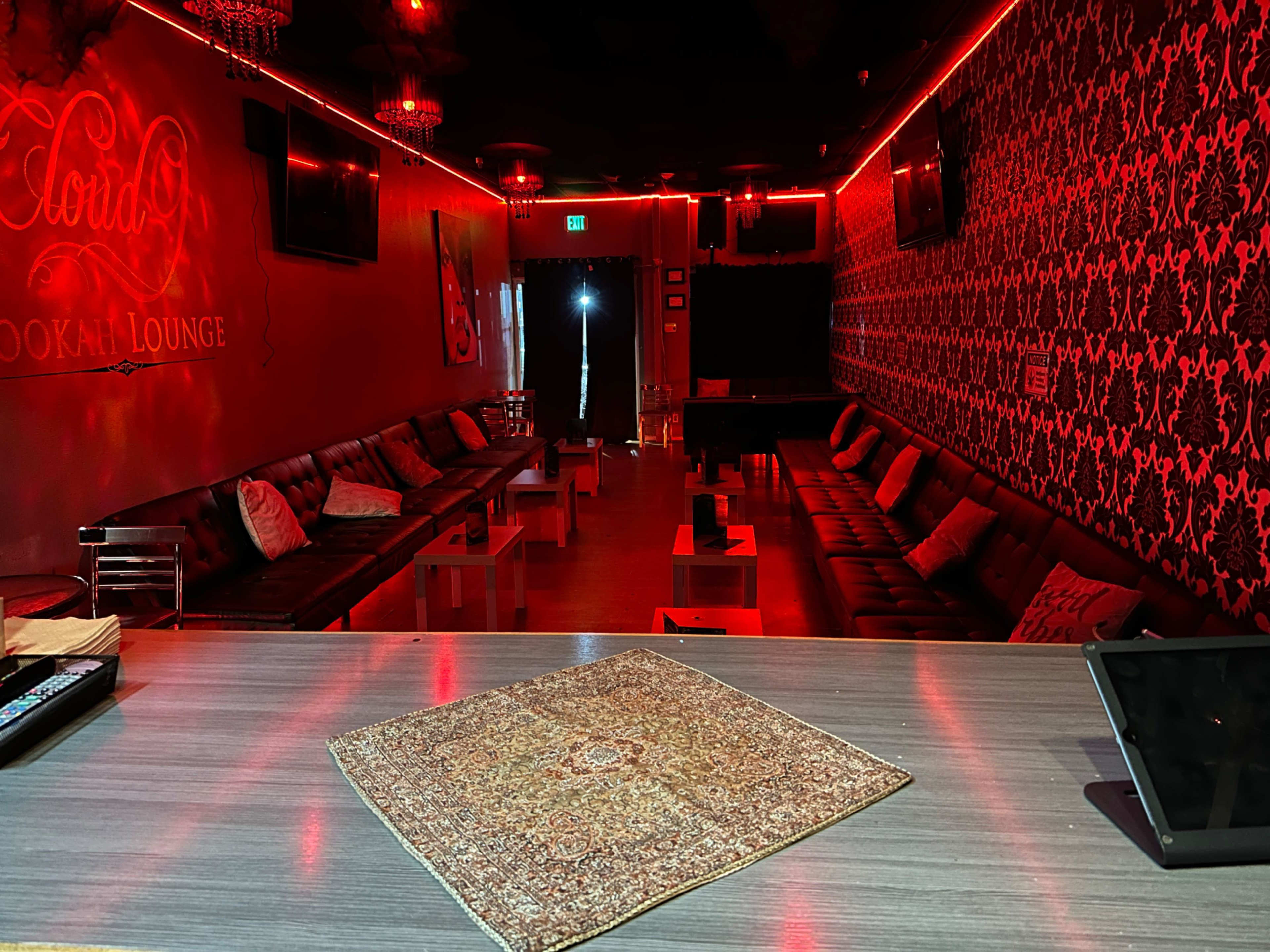 A dimly lit lounge with red lighting, featuring leather seating arranged along the walls and a bar area in the foreground.