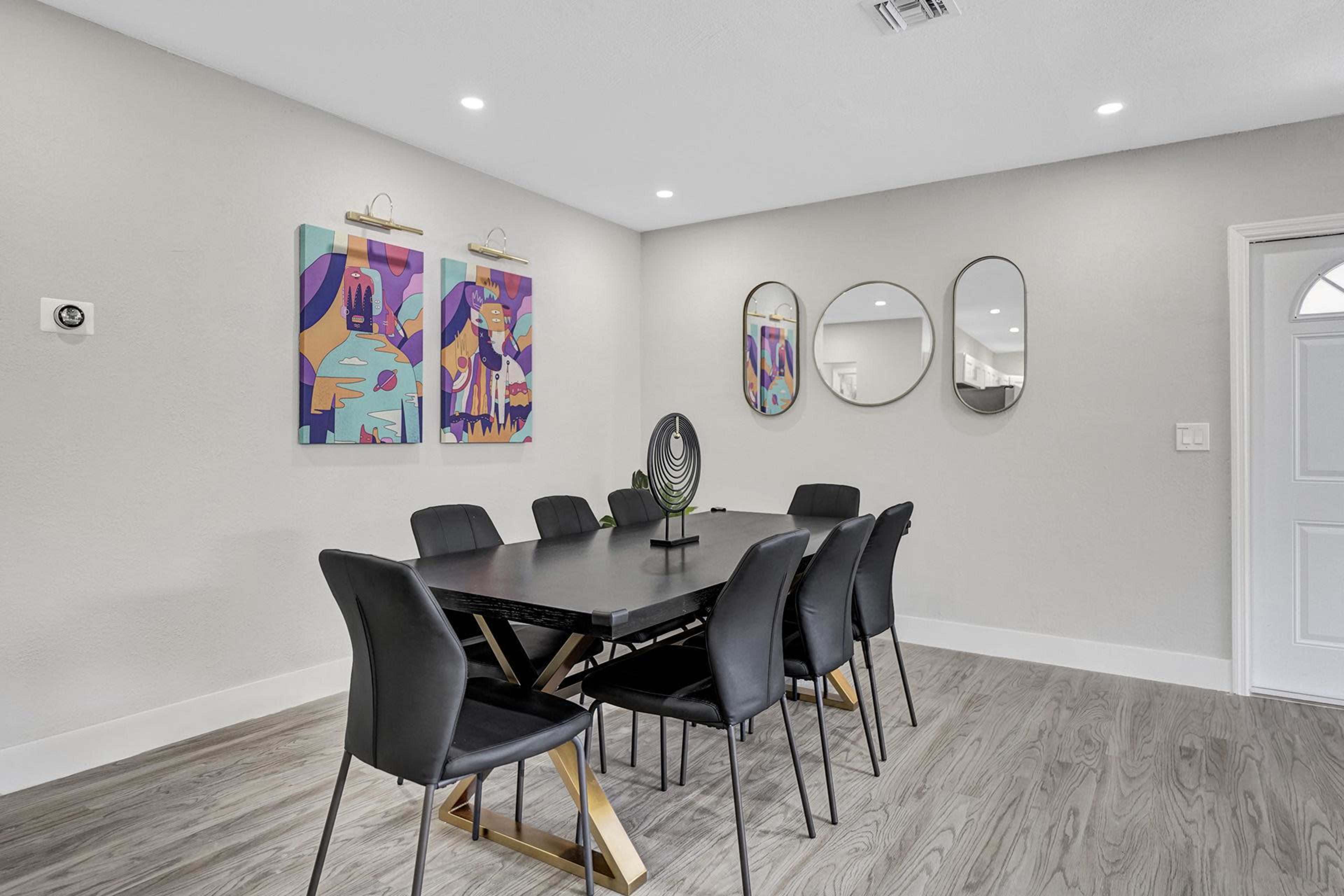 A dining area features a black table surrounded by six chairs, two colorful artworks on the wall, and three round mirrors.