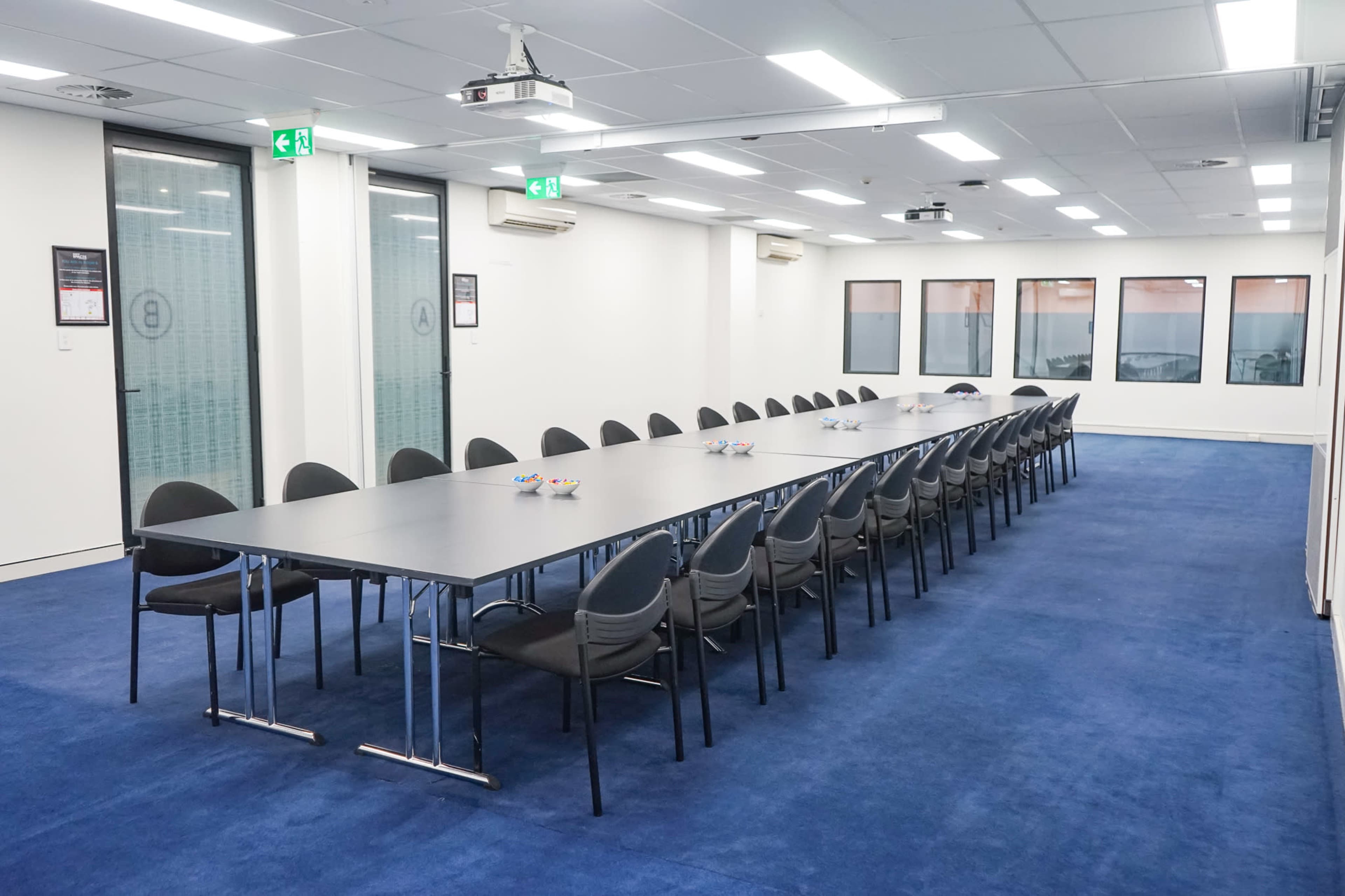 A long, gray conference table surrounded by black chairs is set up in a well-lit meeting room with large windows and blue carpeting.