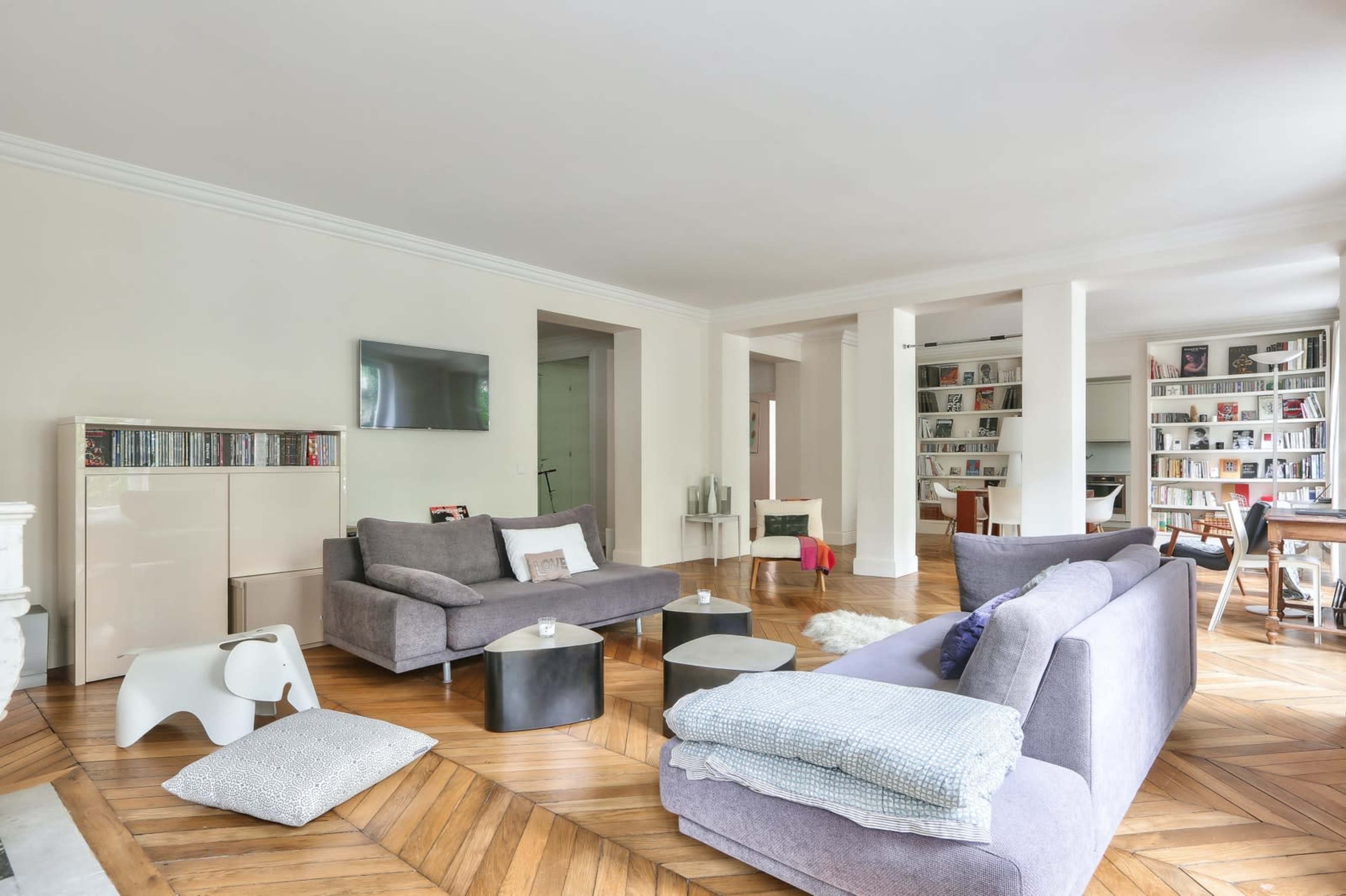 The image shows a spacious living room featuring a mix of modern furniture, hardwood flooring, and shelves filled with books.