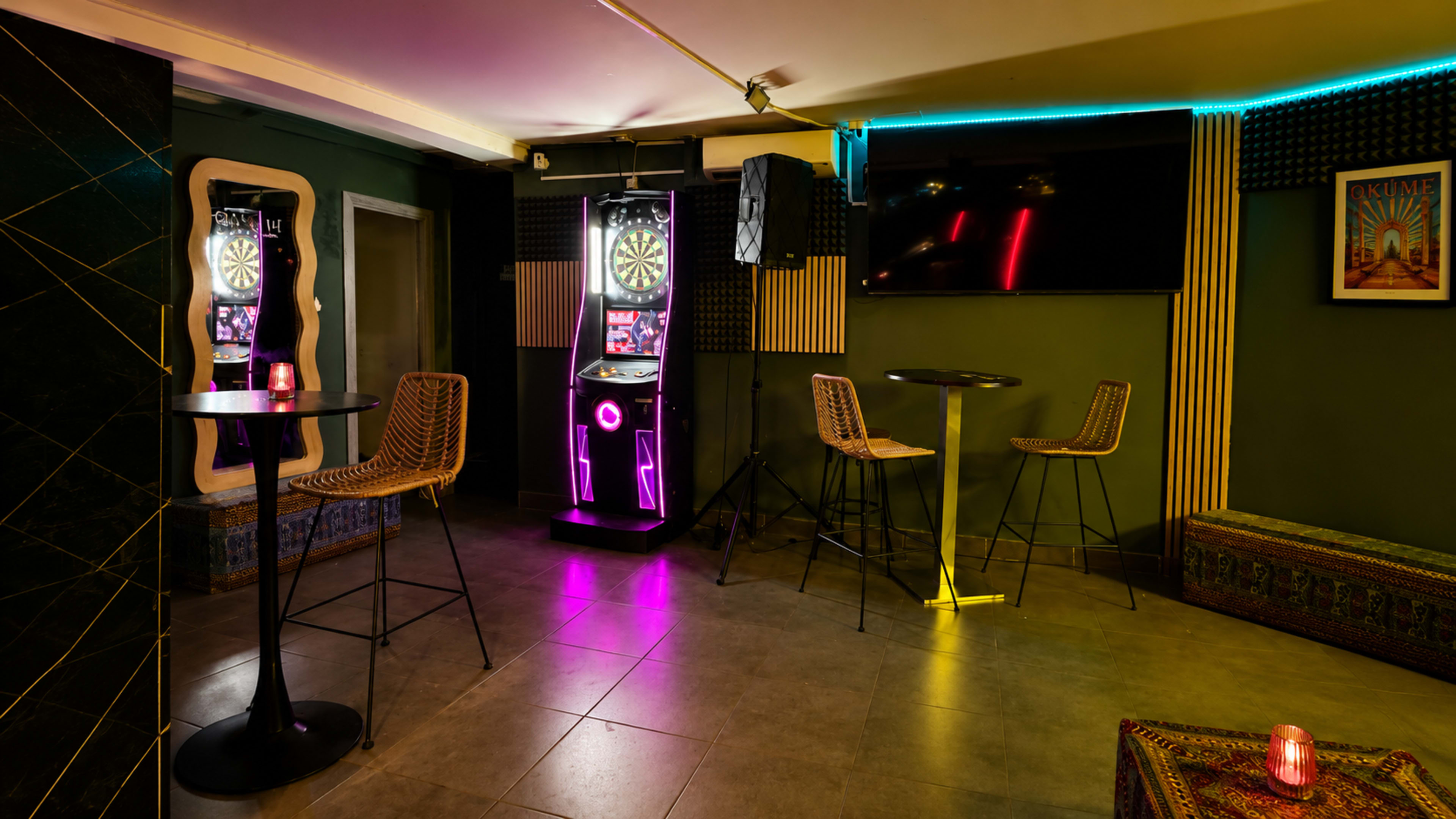 The image shows a modern lounge area with a dartboard machine, bar tables with stools, and ambient lighting.