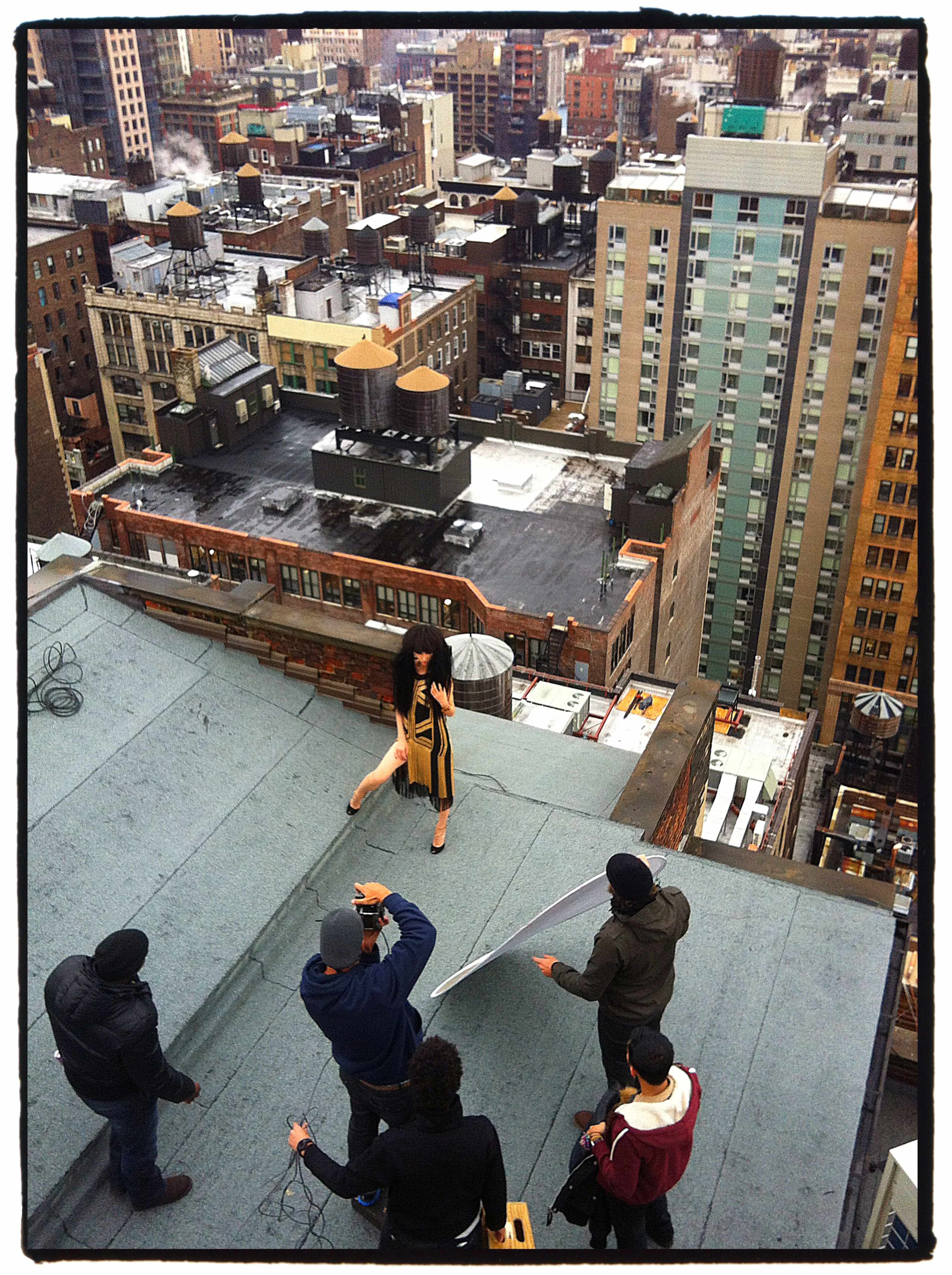 A model poses for a photoshoot on a rooftop in an urban setting while a small crew adjusts lighting and equipment.