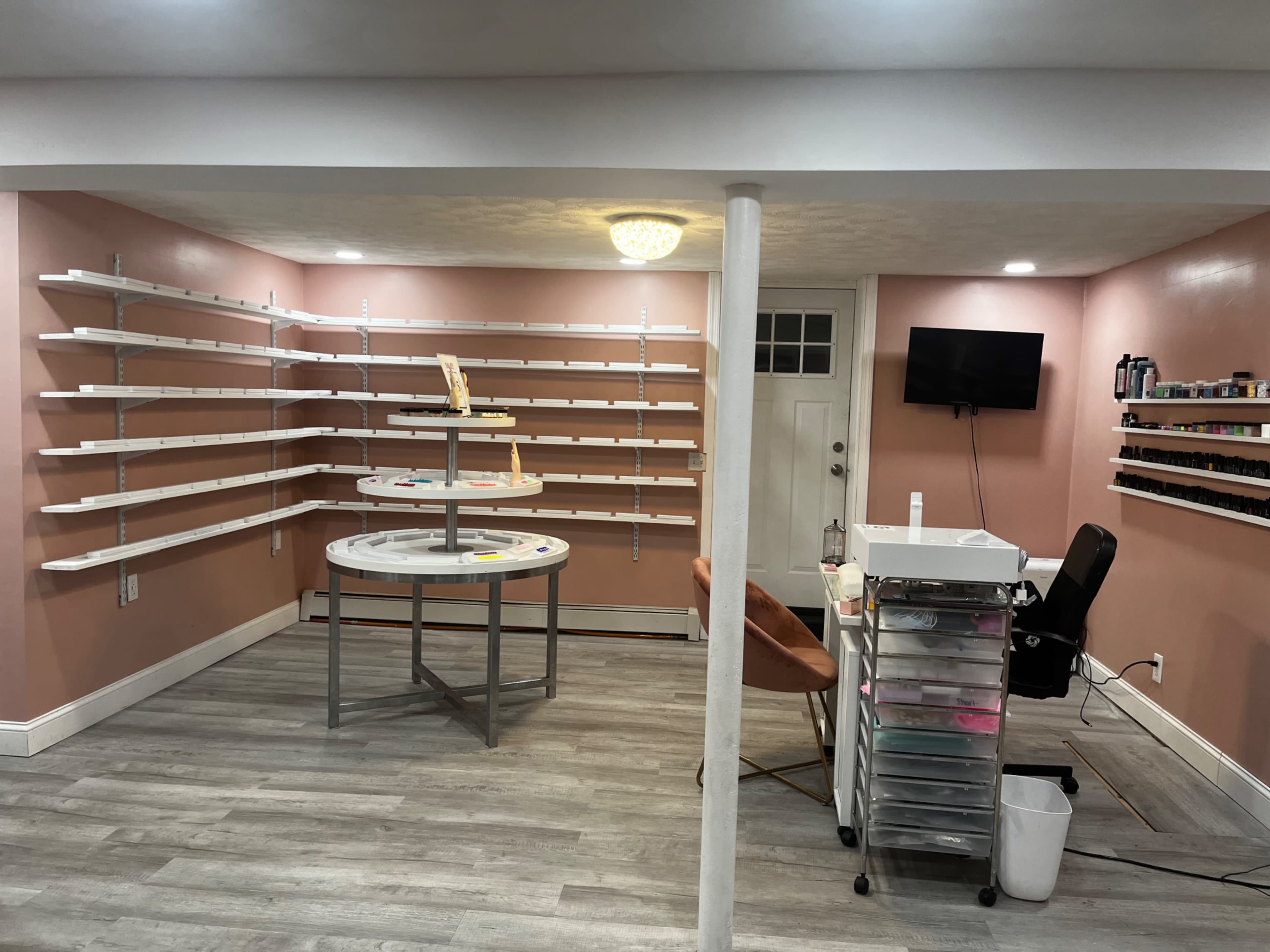 A well-lit room with pink walls, featuring white shelves on either side, a circular display table in the center, and a desk with a chair against the wall.
