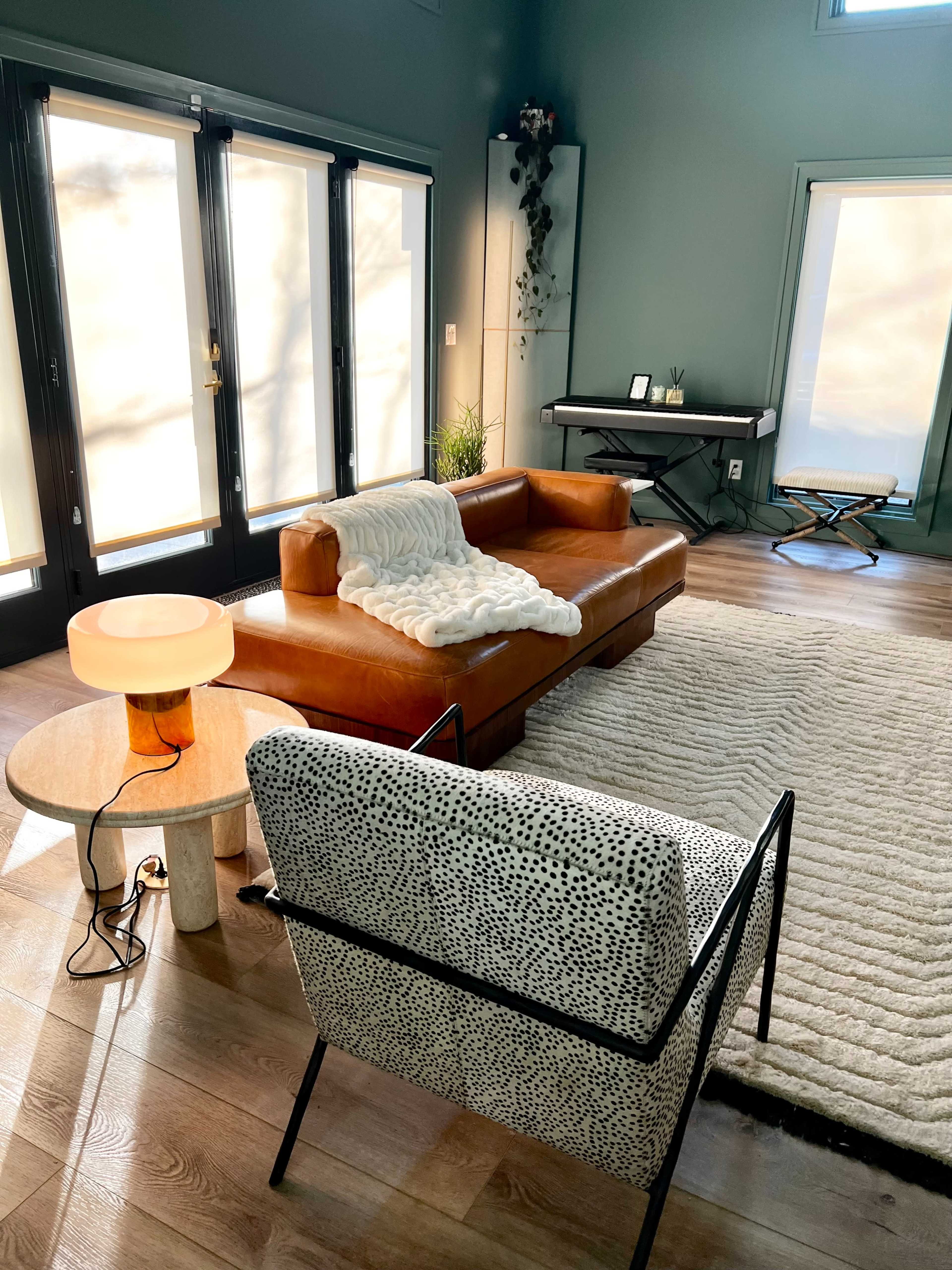 The image shows a modern living room with a brown leather sofa, a round coffee table, a patterned armchair, and large windows allowing natural light to fill the space.