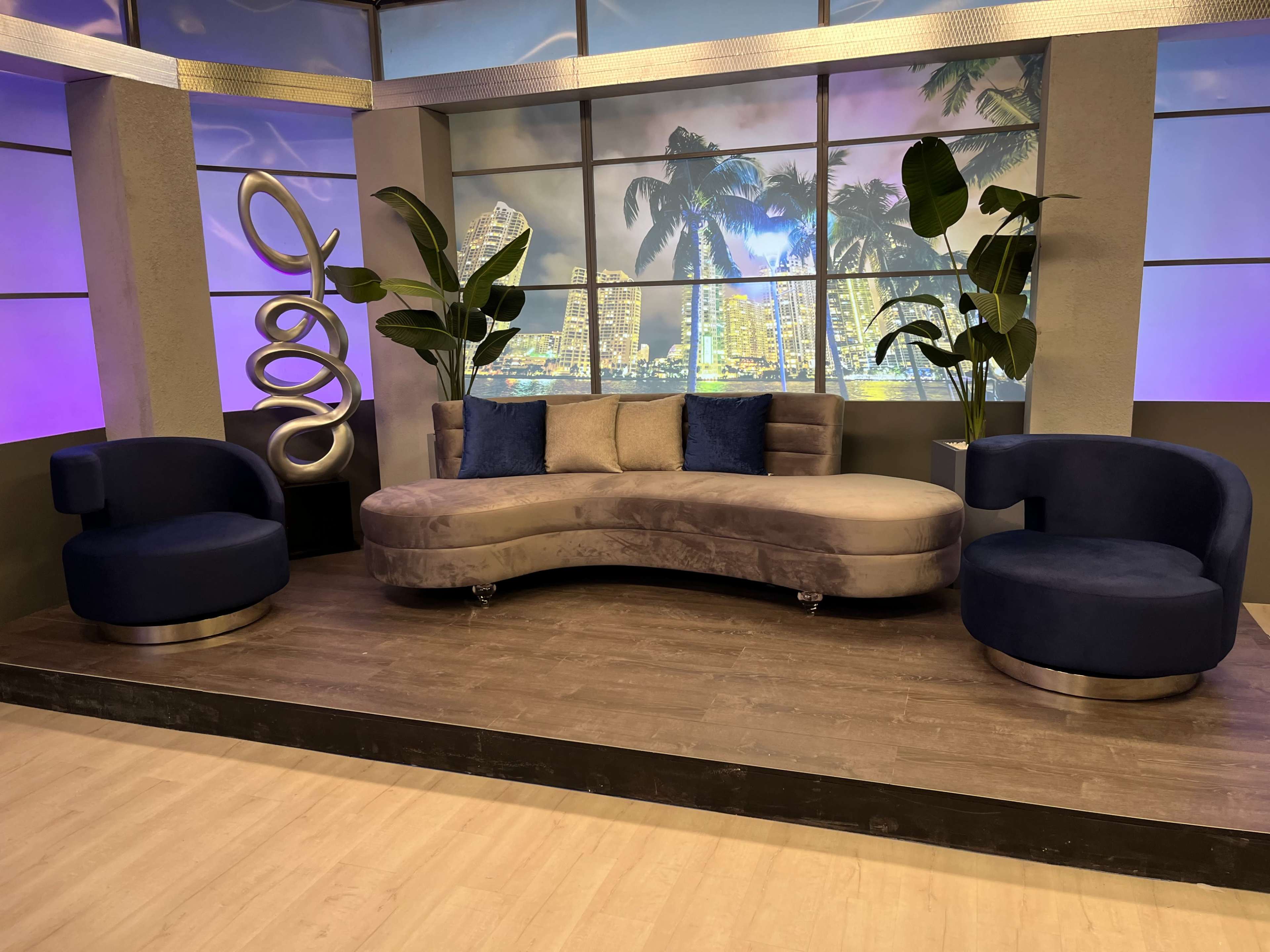 A modern television set featuring a curved gray sofa flanked by two dark blue chairs, with large potted plants and a city skyline backdrop.