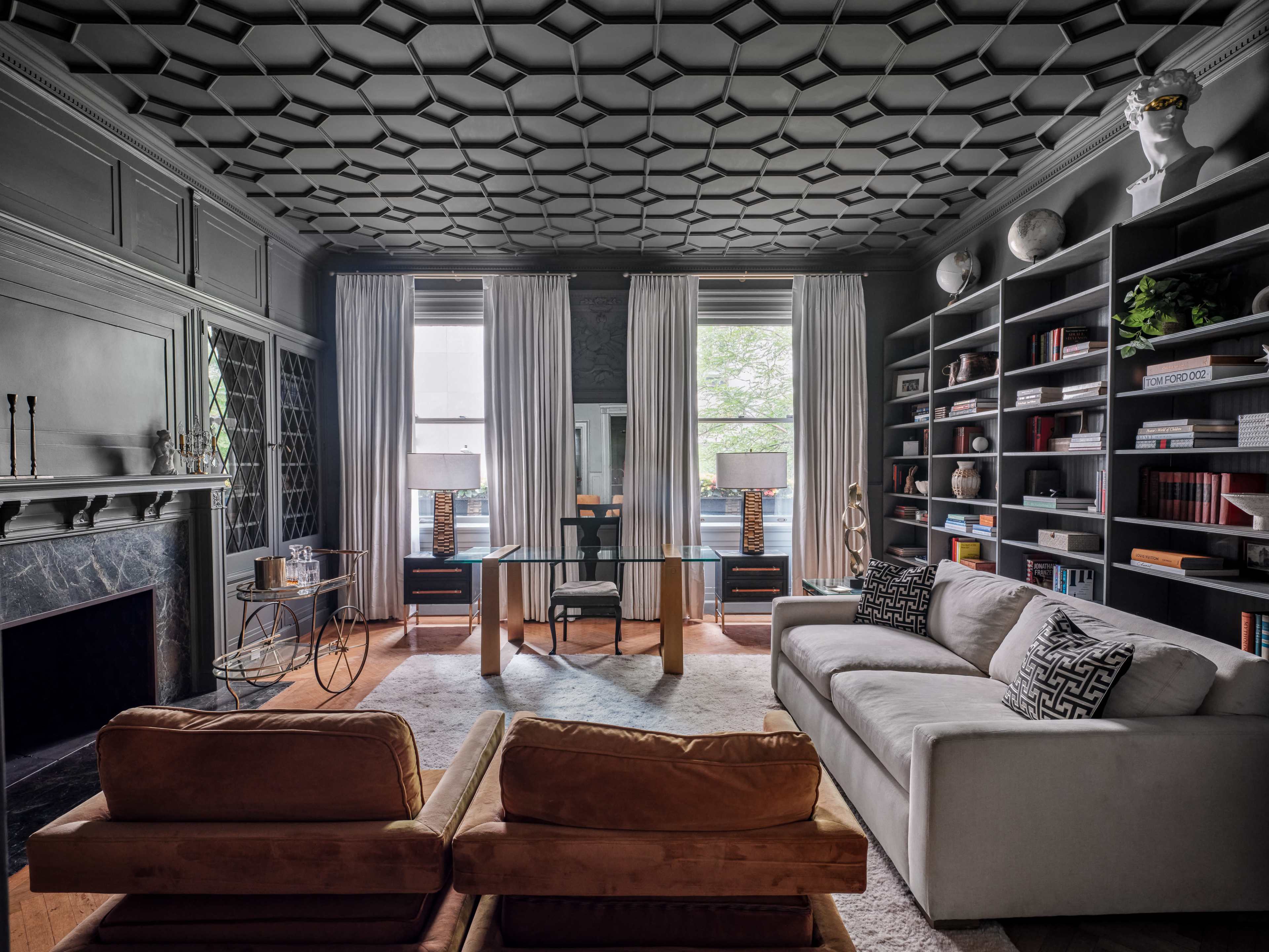 A sophisticated living room with a patterned ceiling, a cozy sofa, a glass-top desk flanked by large windows, and a bookshelf filled with books.