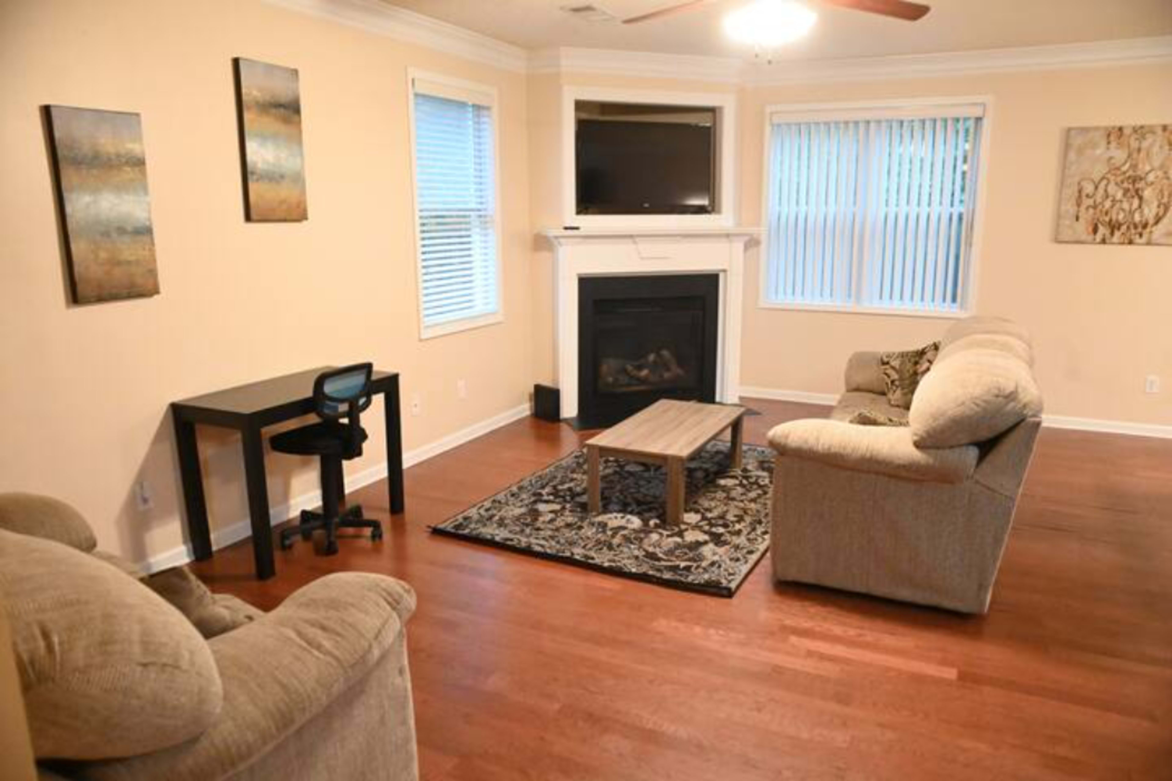 The room features a fireplace, a television above it, a coffee table, a desk with a chair, and two sofas arranged around a patterned rug on wooden flooring.