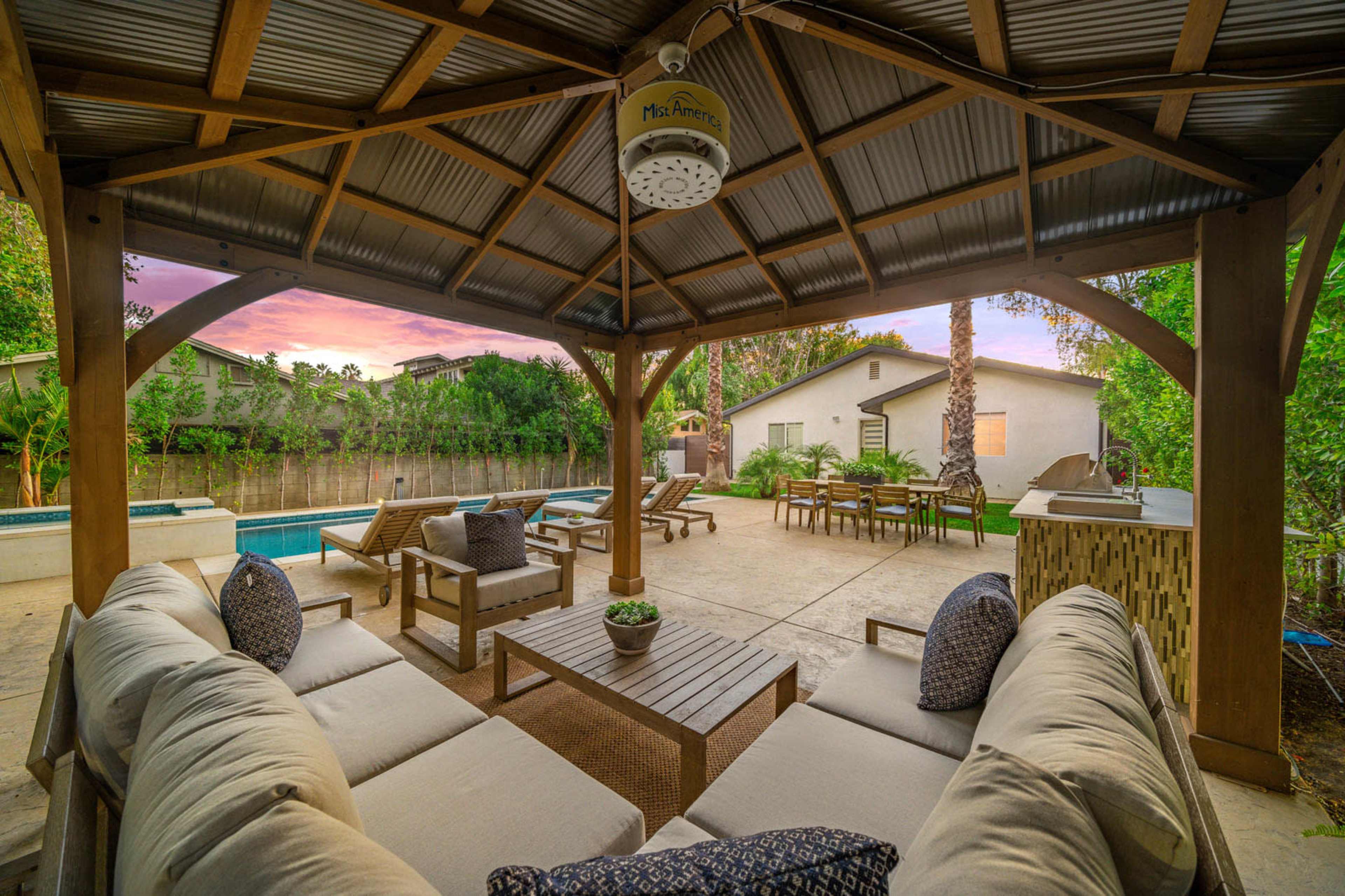 The image shows a spacious outdoor patio with a gazebo, comfortable seating, and a pool area surrounded by greenery and a house in the background.