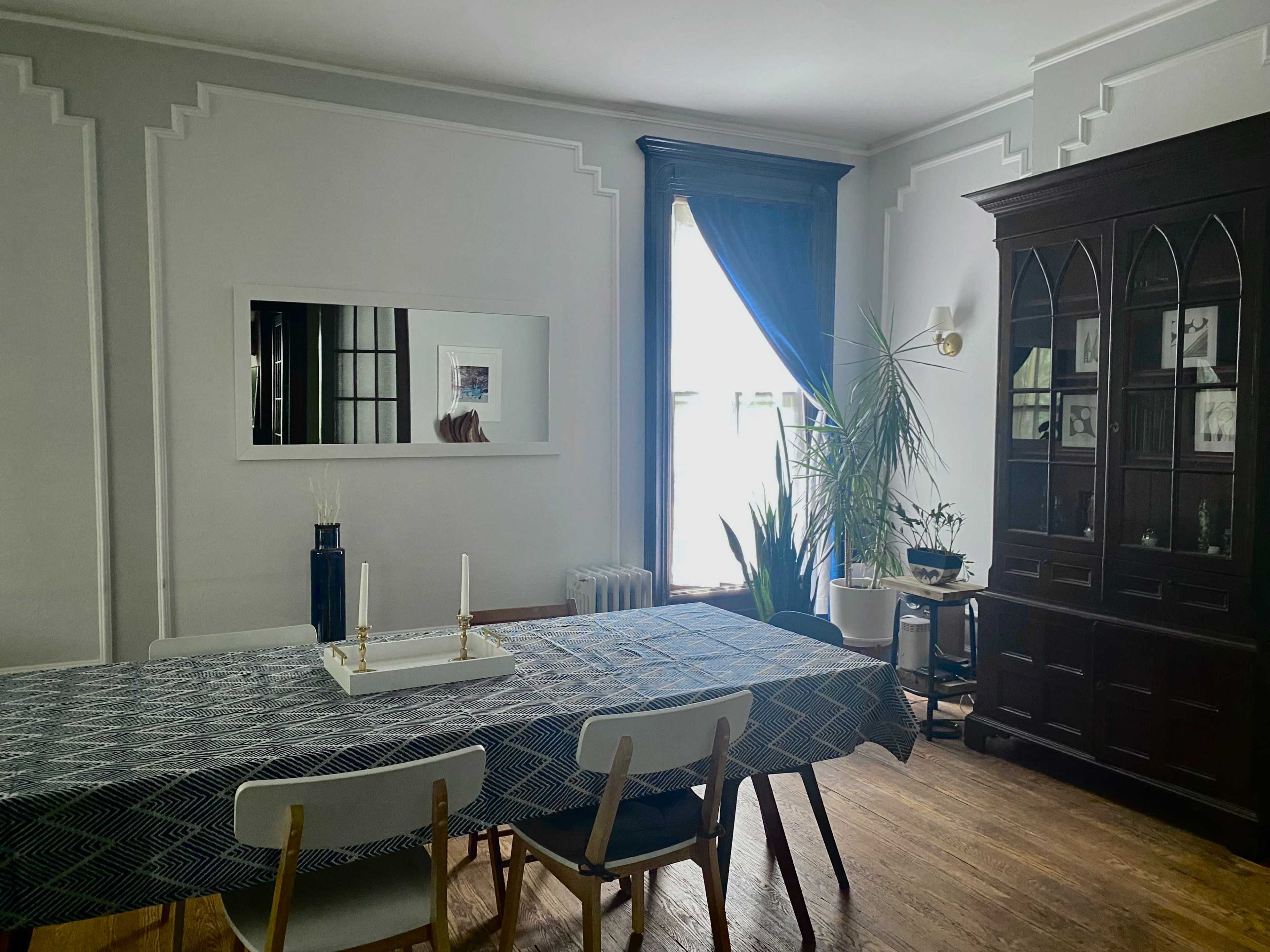 A dining room features a long table set with a blue patterned tablecloth, surrounded by white chairs, and is adorned with a tall mirror, a plant, and a dark wooden cabinet against the wall.
