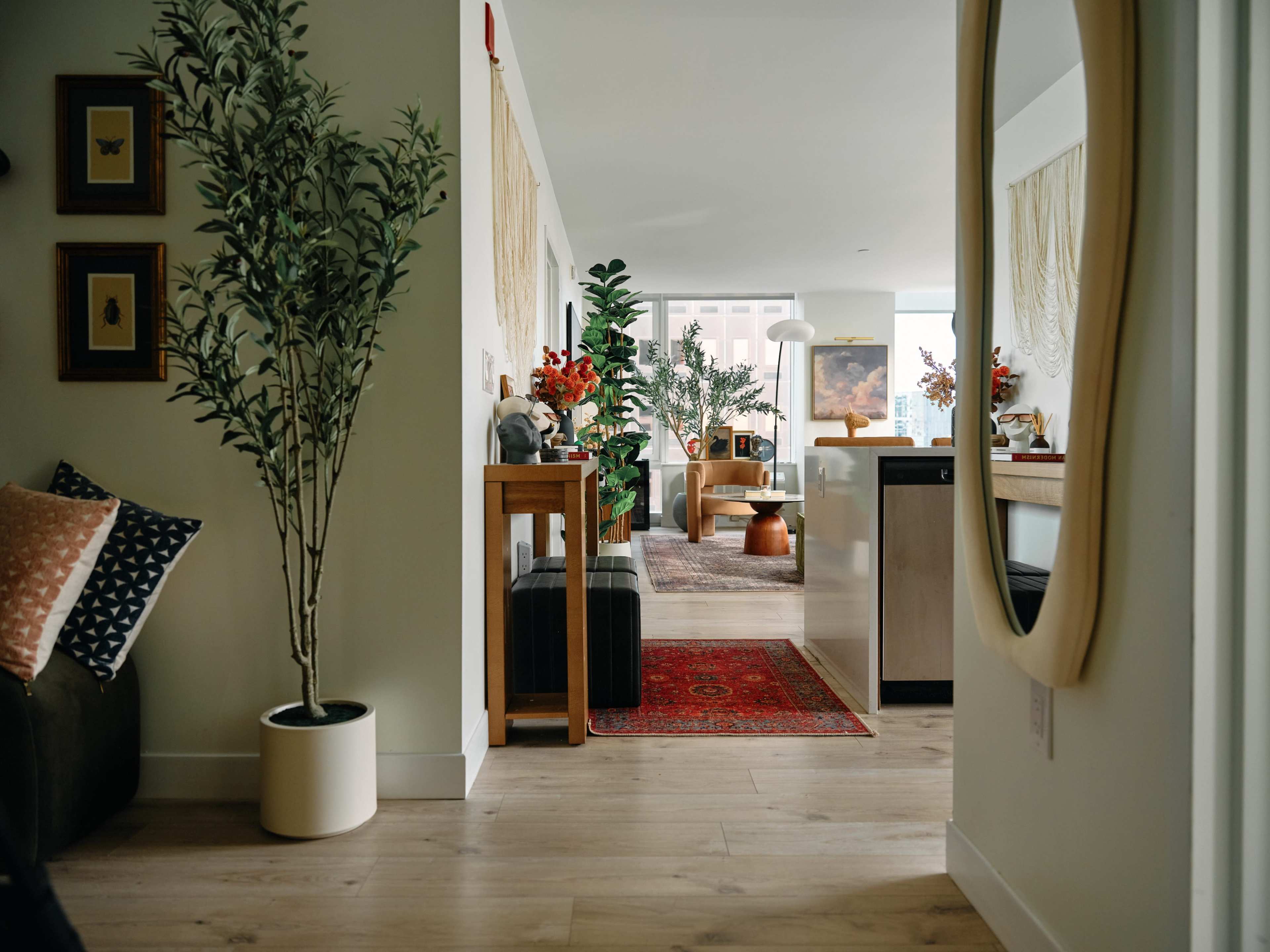 A cozy, modern interior features a hallway leading to a well-decorated living area with plants, artwork, and furniture arranged around a central rug.