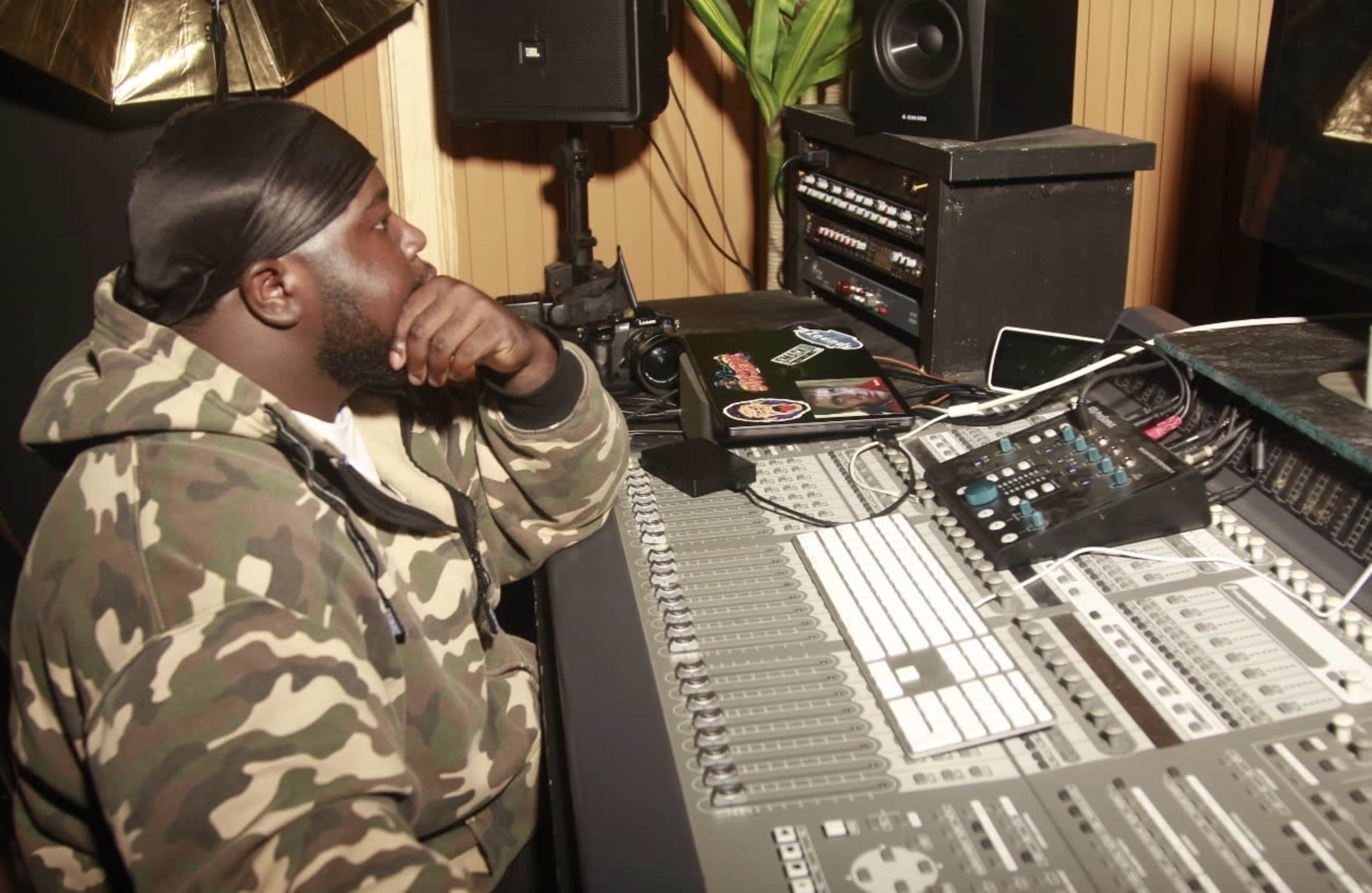 A person in a camouflage hoodie sits at a recording studio mixing console, focused on the equipment in front of them.