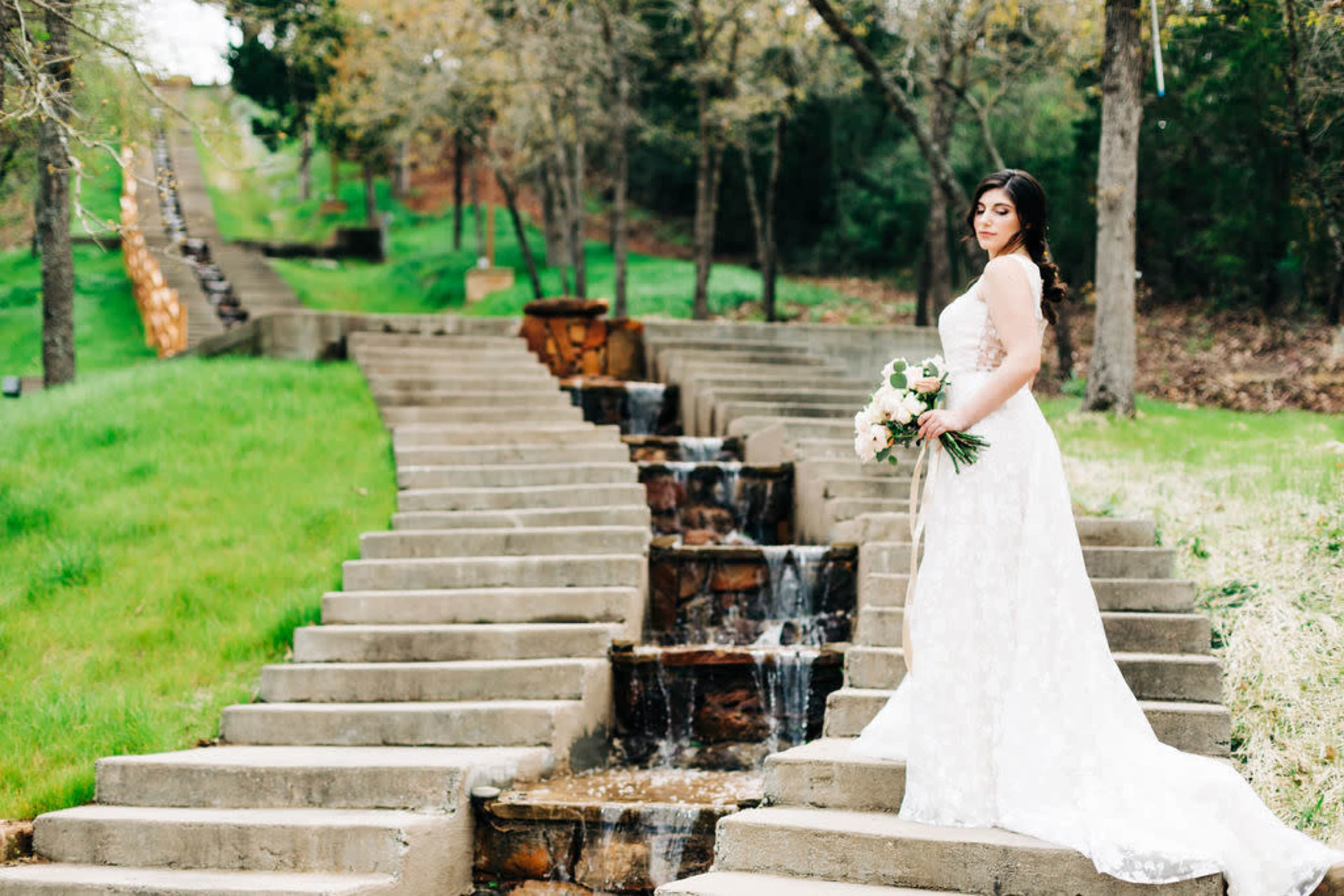 A bride in a white lace dress stands on stone steps beside a small waterfall in a green, landscaped area.