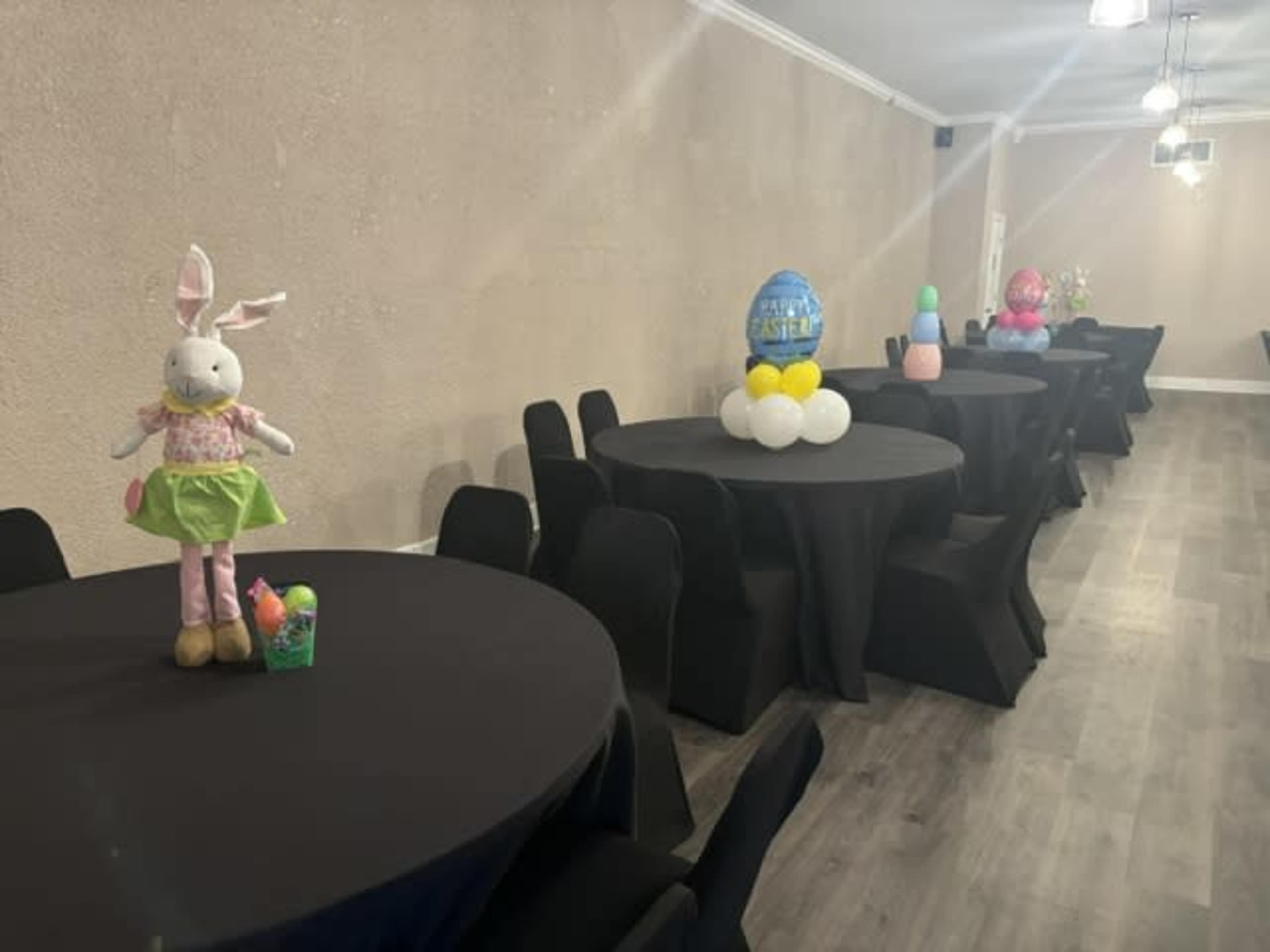 The image shows a decorated event space with round tables covered in black tablecloths, featuring colorful Easter-themed decorations.