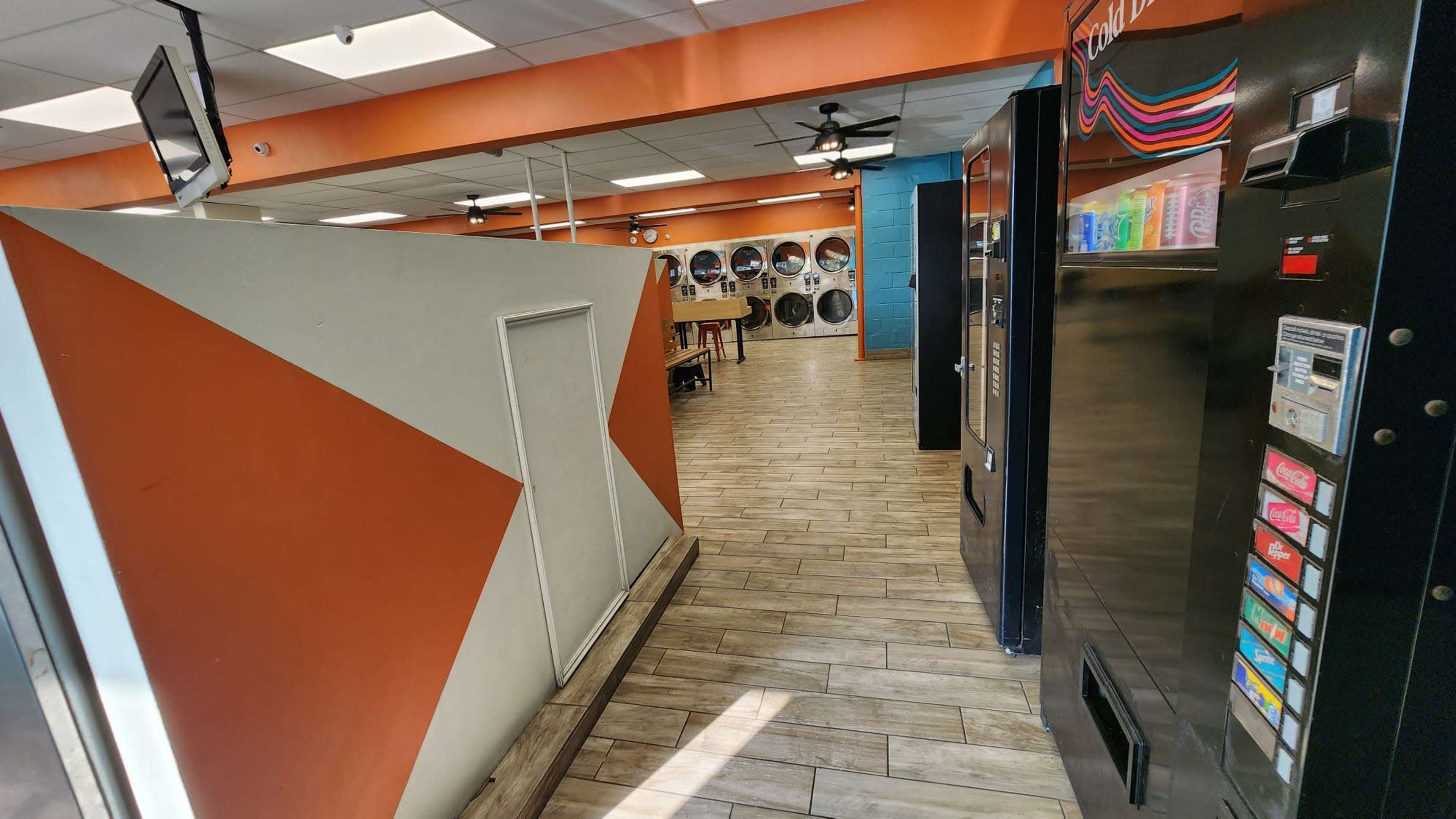 A narrow corridor in a laundromat with vending machines on the right and washing machines visible in a designated area ahead.