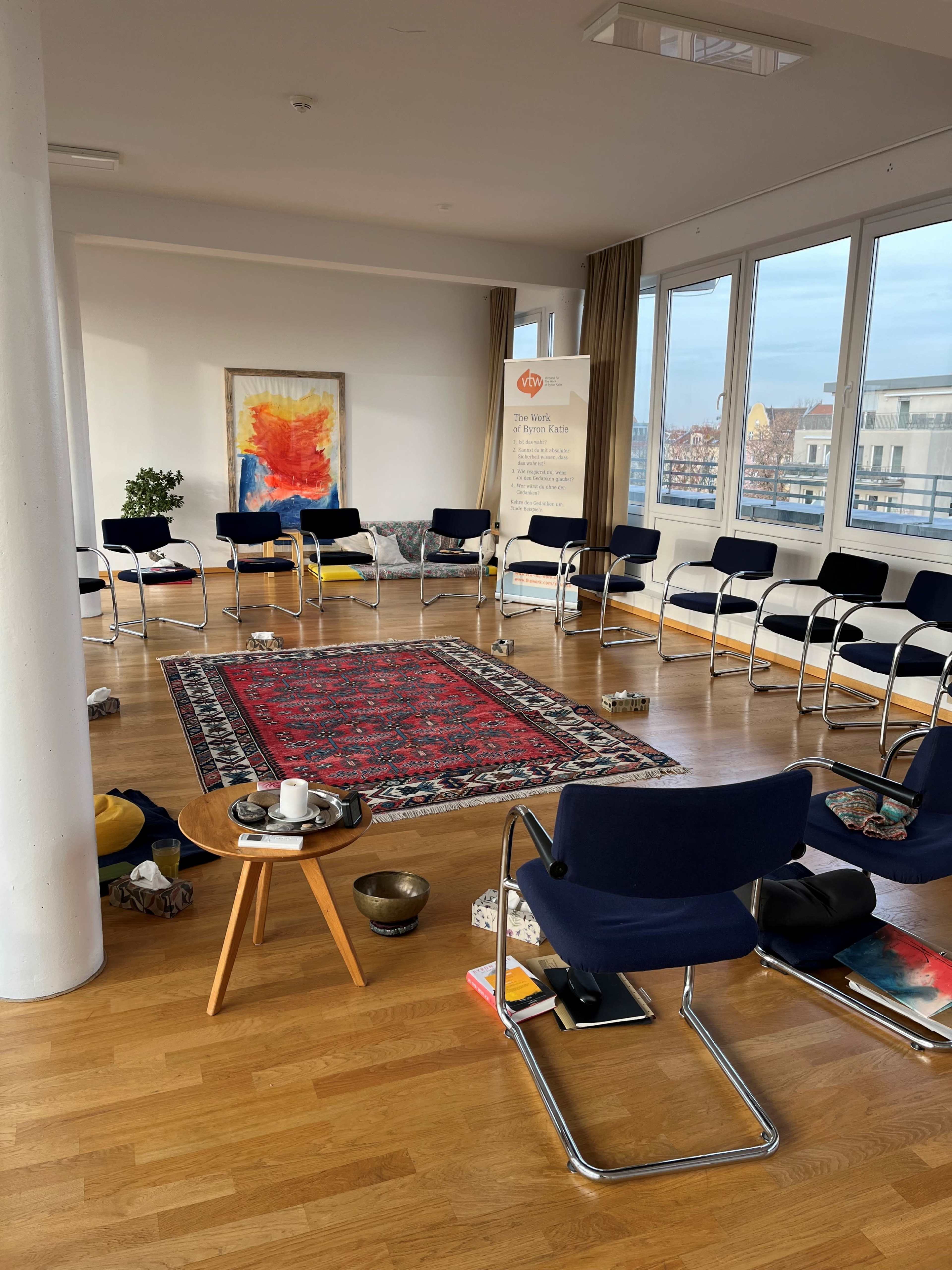 A circular seating arrangement with chairs surrounds a Persian rug, while various items are scattered across the wooden floor in a well-lit room.