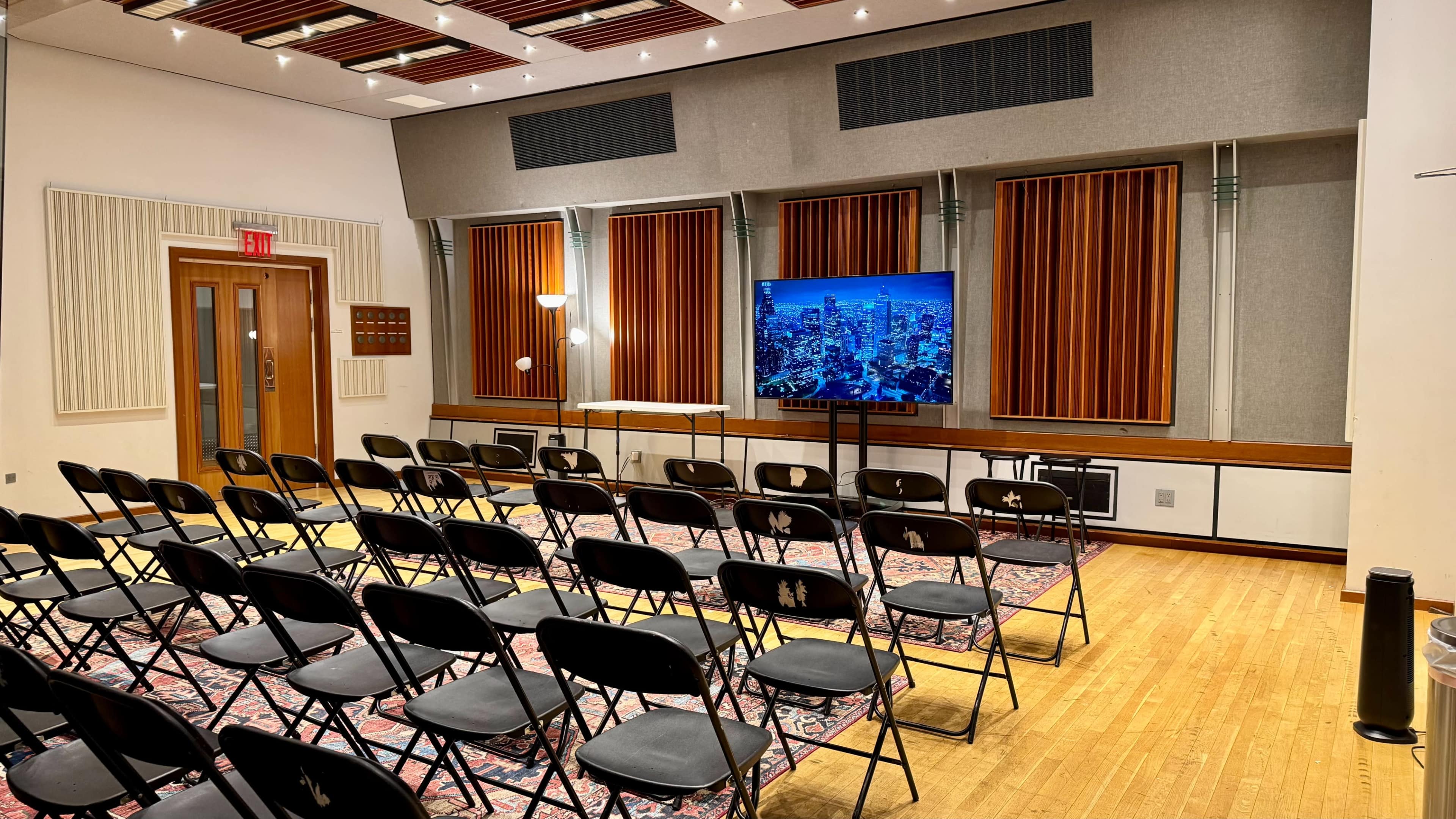 The image shows a seminar room with rows of black chairs facing a wall-mounted screen displaying a city skyline, flanked by wooden acoustic panels.