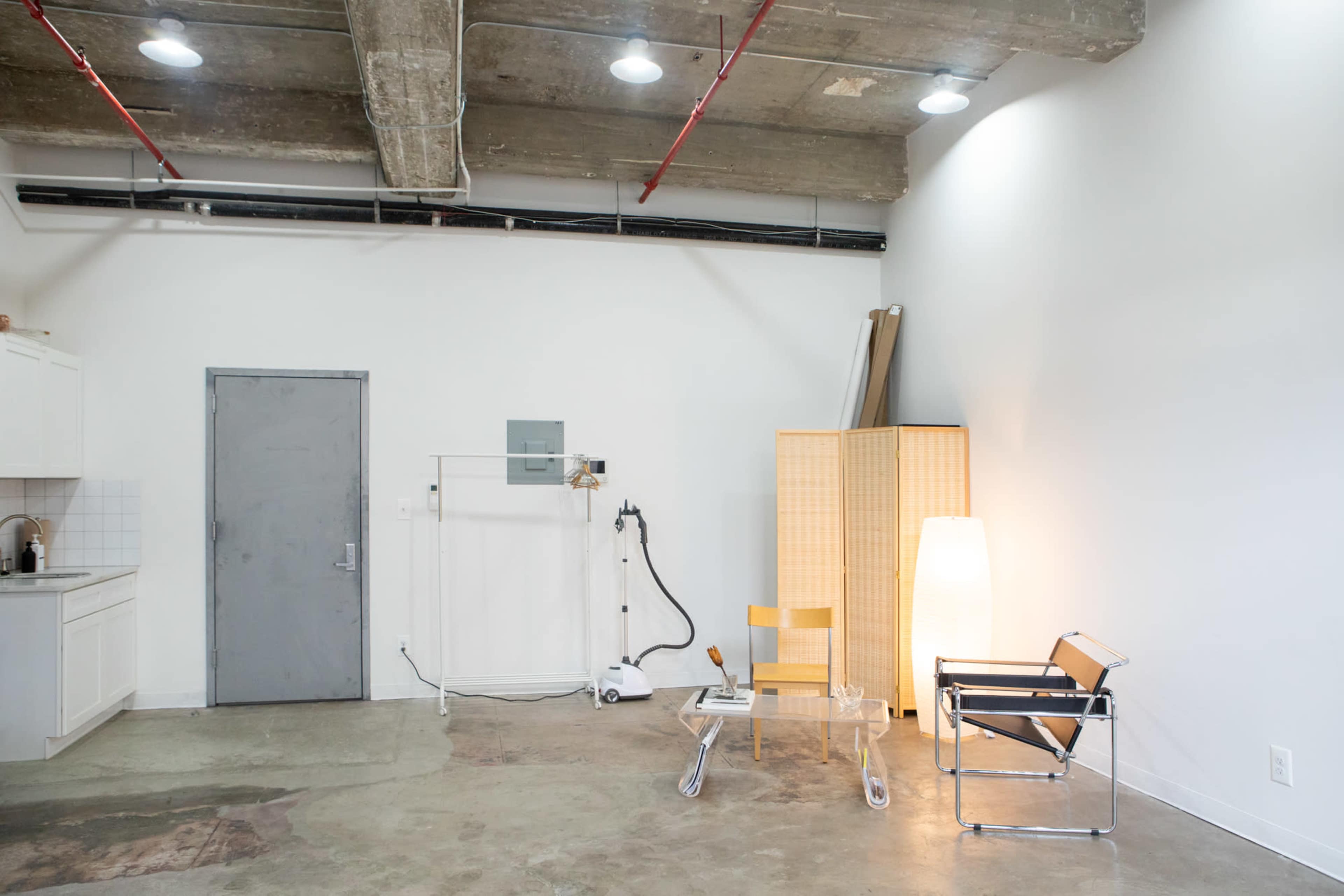 A minimalist room with a kitchen area, a door, a folding screen, a lamp, and two chairs on a concrete floor.