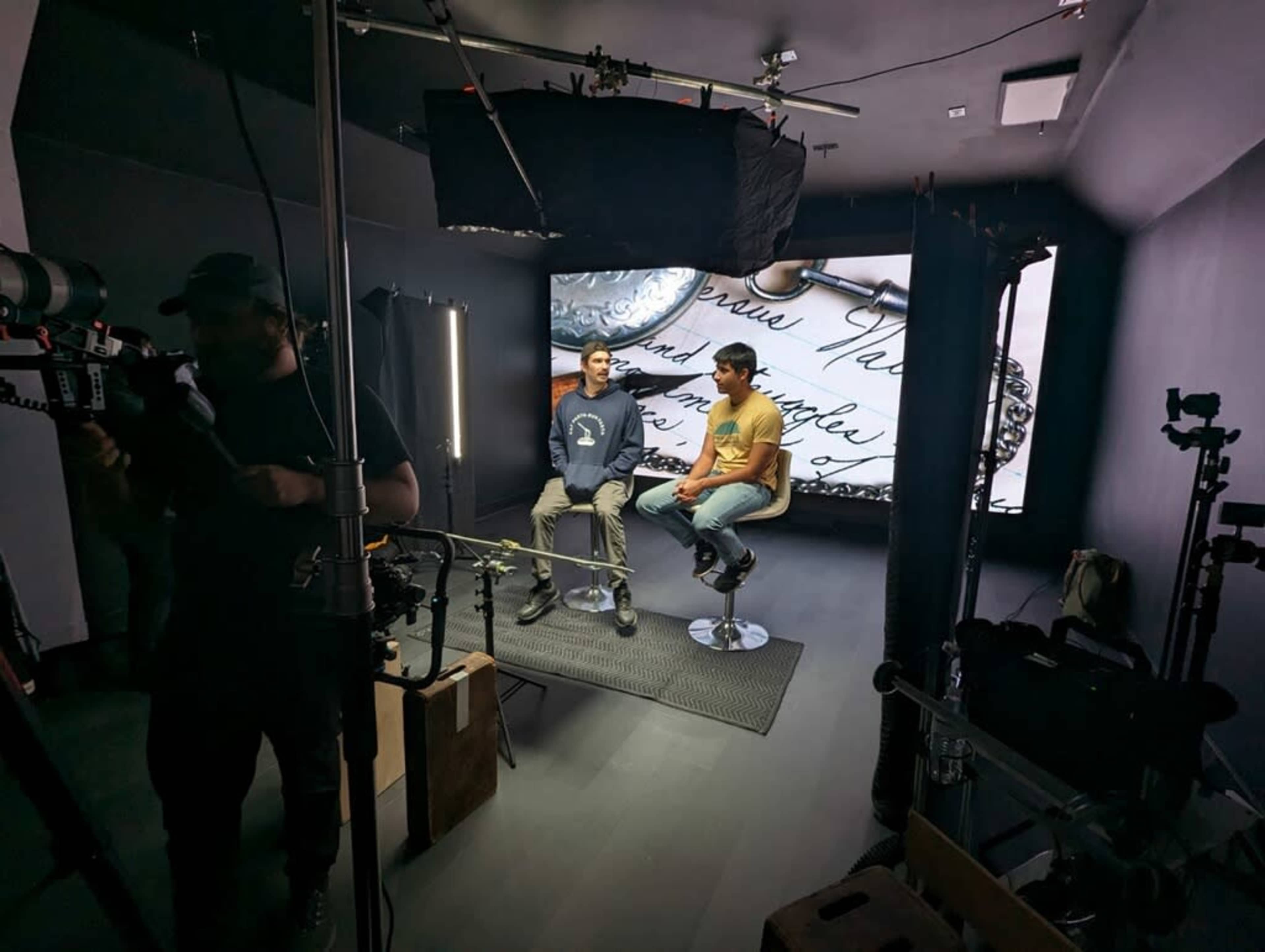 Two people sit on stools in a studio setting, with a large screen displaying handwritten text in the background and various film equipment surrounding them.