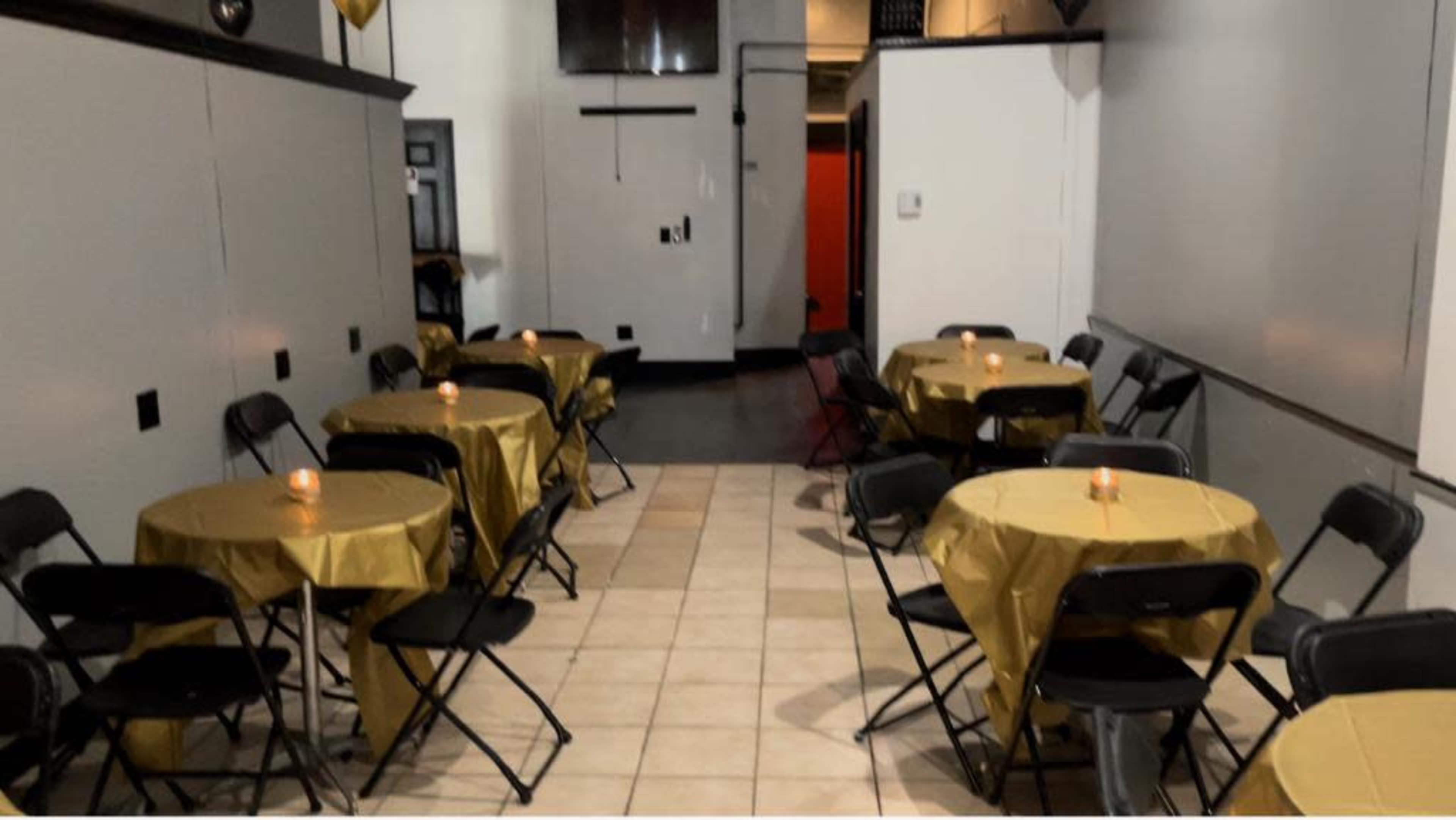 An interior space with several round tables covered in gold tablecloths, each accompanied by black chairs and lit candles, creating a simple dining setup.