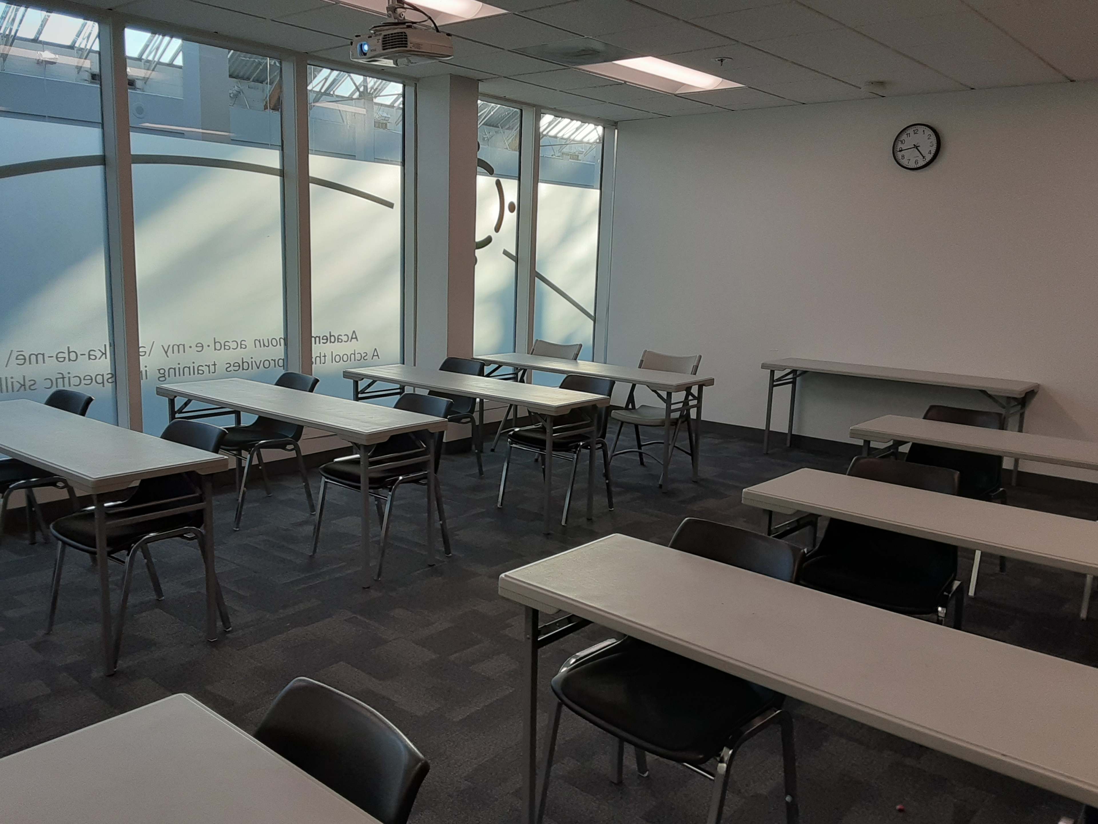 A classroom with several rows of tables and chairs, a wall clock, and large windows allowing natural light to enter.