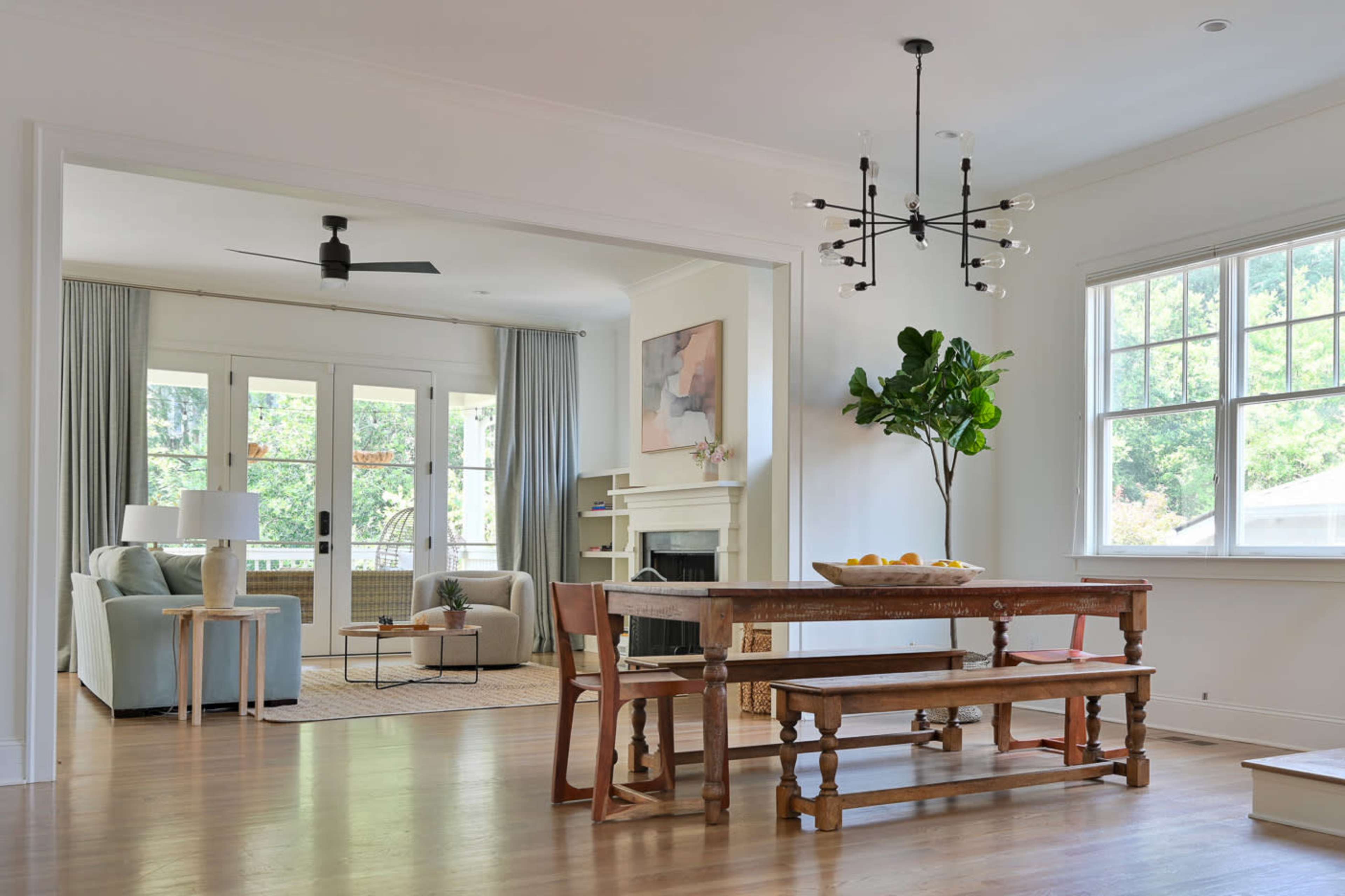 A modern dining area with a wooden table and benches, leading into a bright living space that features a fireplace, large windows, and indoor plants.