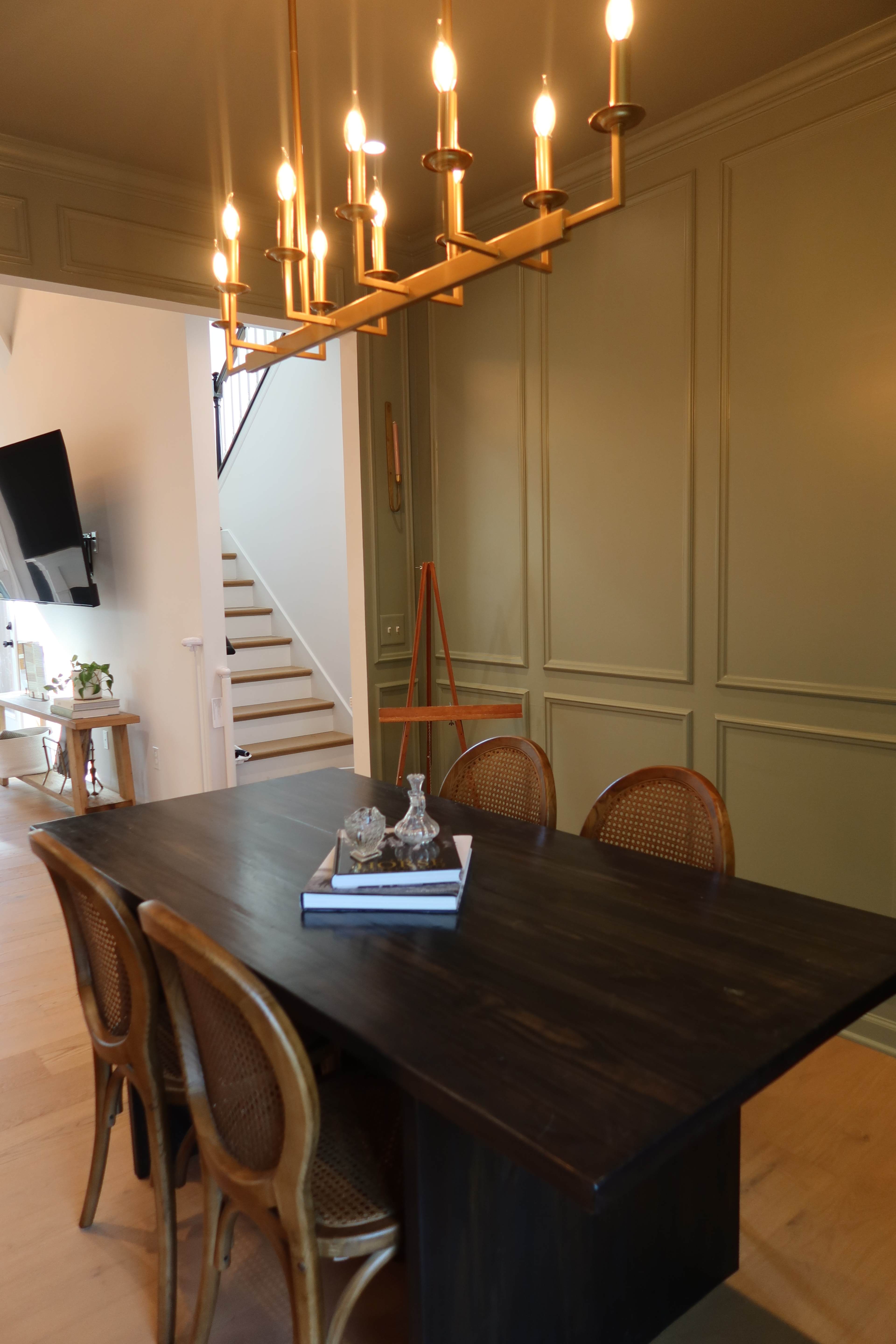 A wooden dining table with four rattan chairs is positioned under a modern chandelier in a well-lit room with green paneled walls.