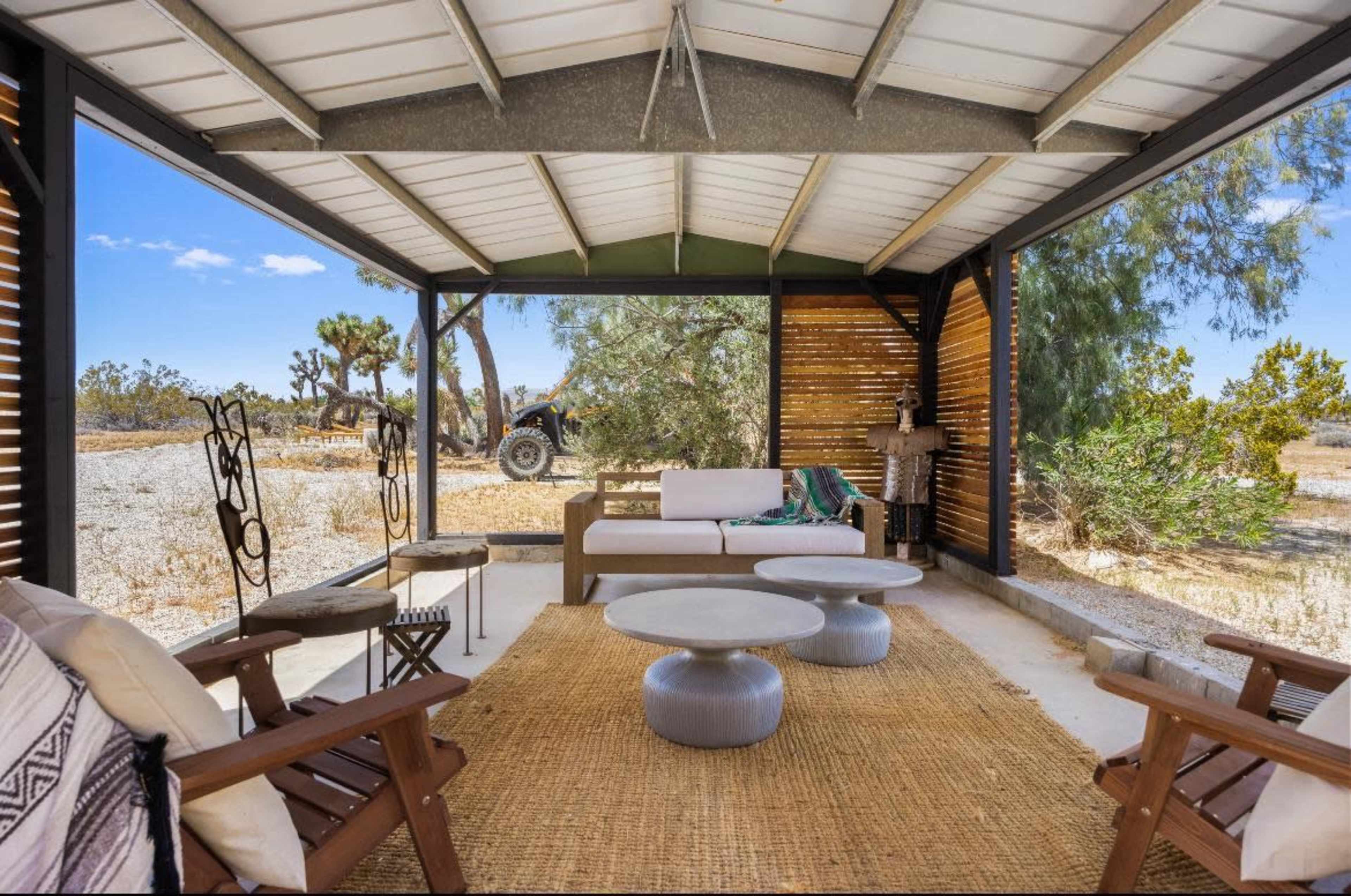 A covered outdoor seating area with a sofa, coffee tables, and a view of a landscape featuring a tractor and sparse vegetation.