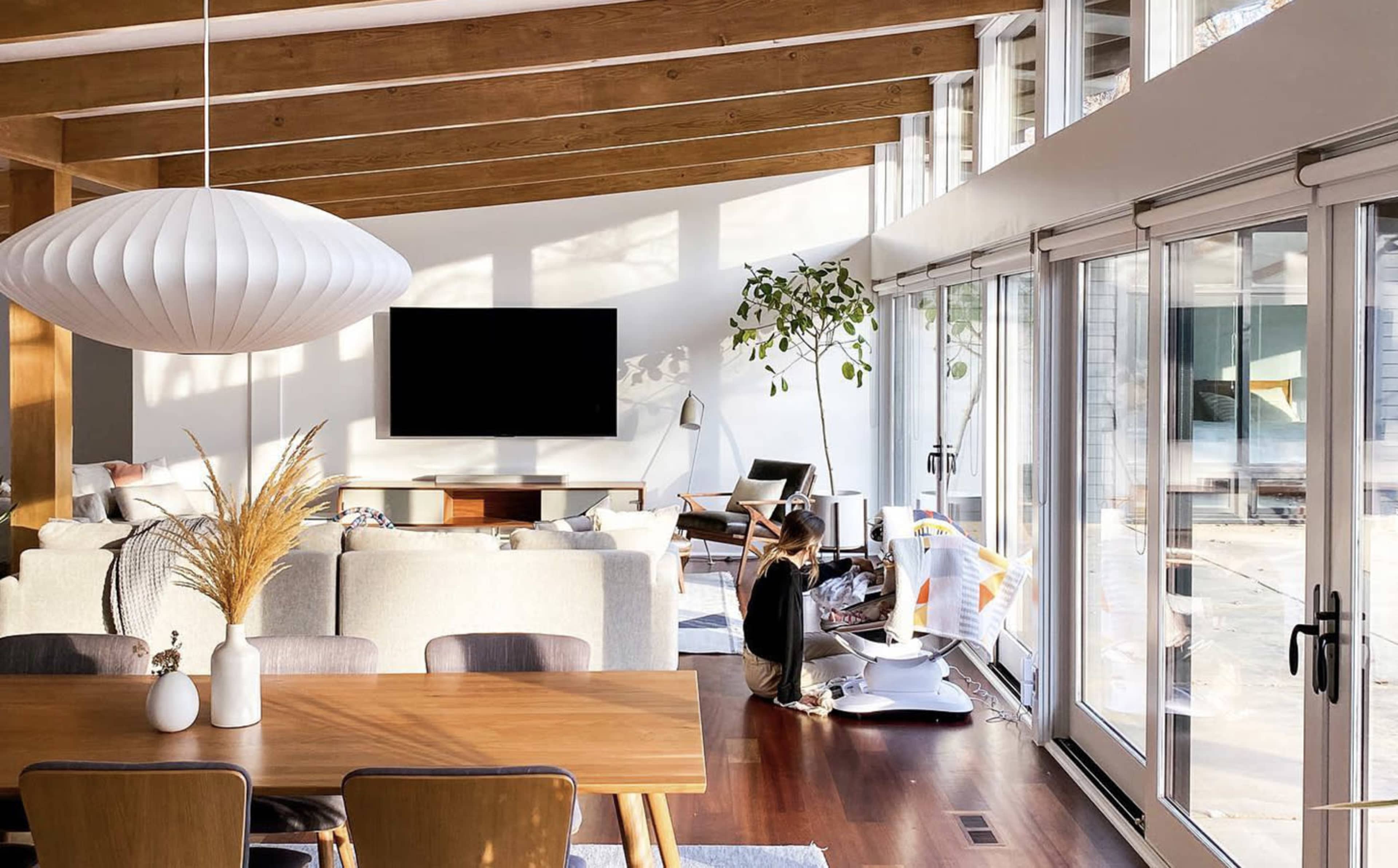 A woman sits on the floor near a modern dining area in a spacious room filled with natural light and large windows, with a television mounted on the wall.
