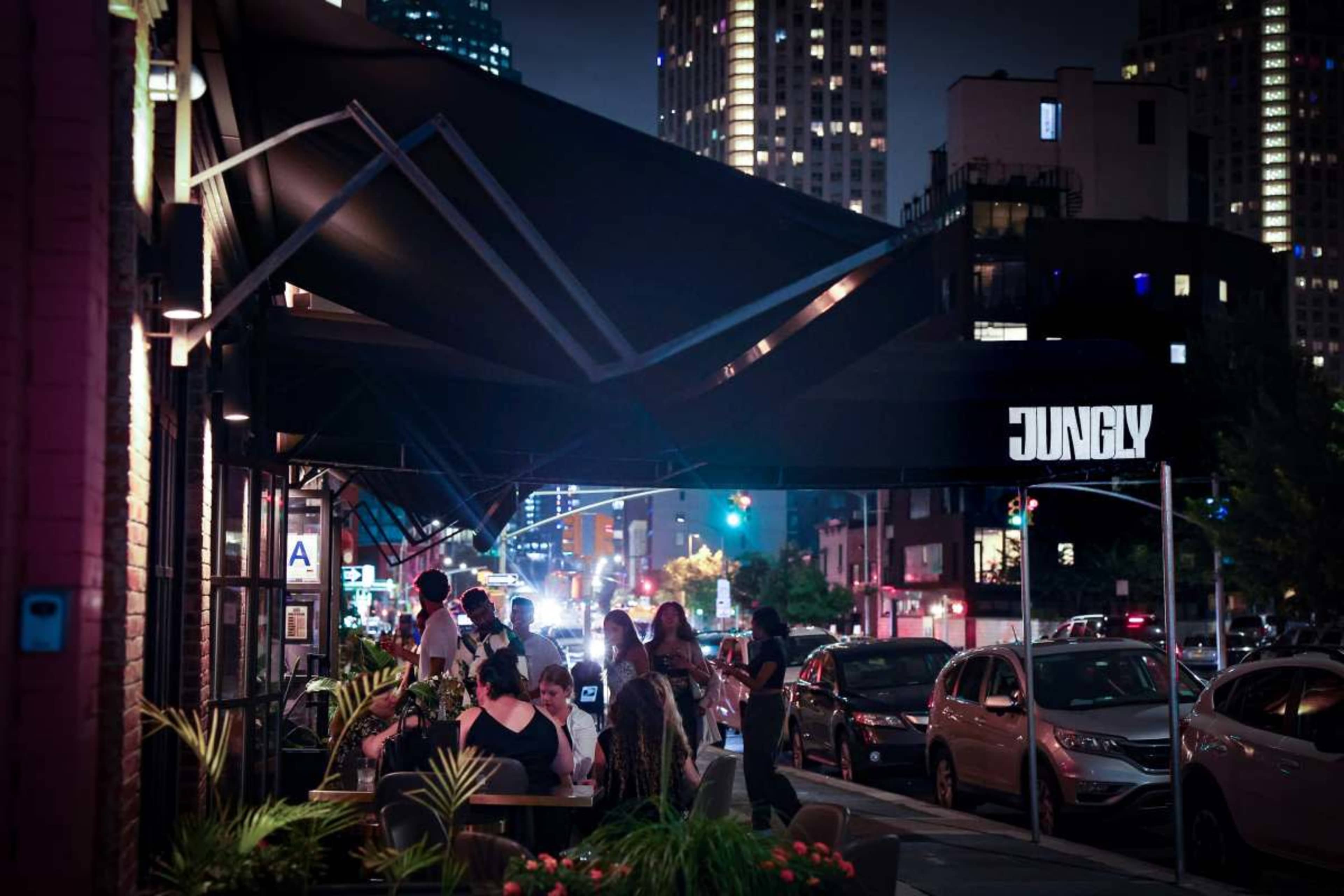 A busy outdoor dining area is illuminated by city lights at night, with people gathered around tables near a restaurant called "Jungly."