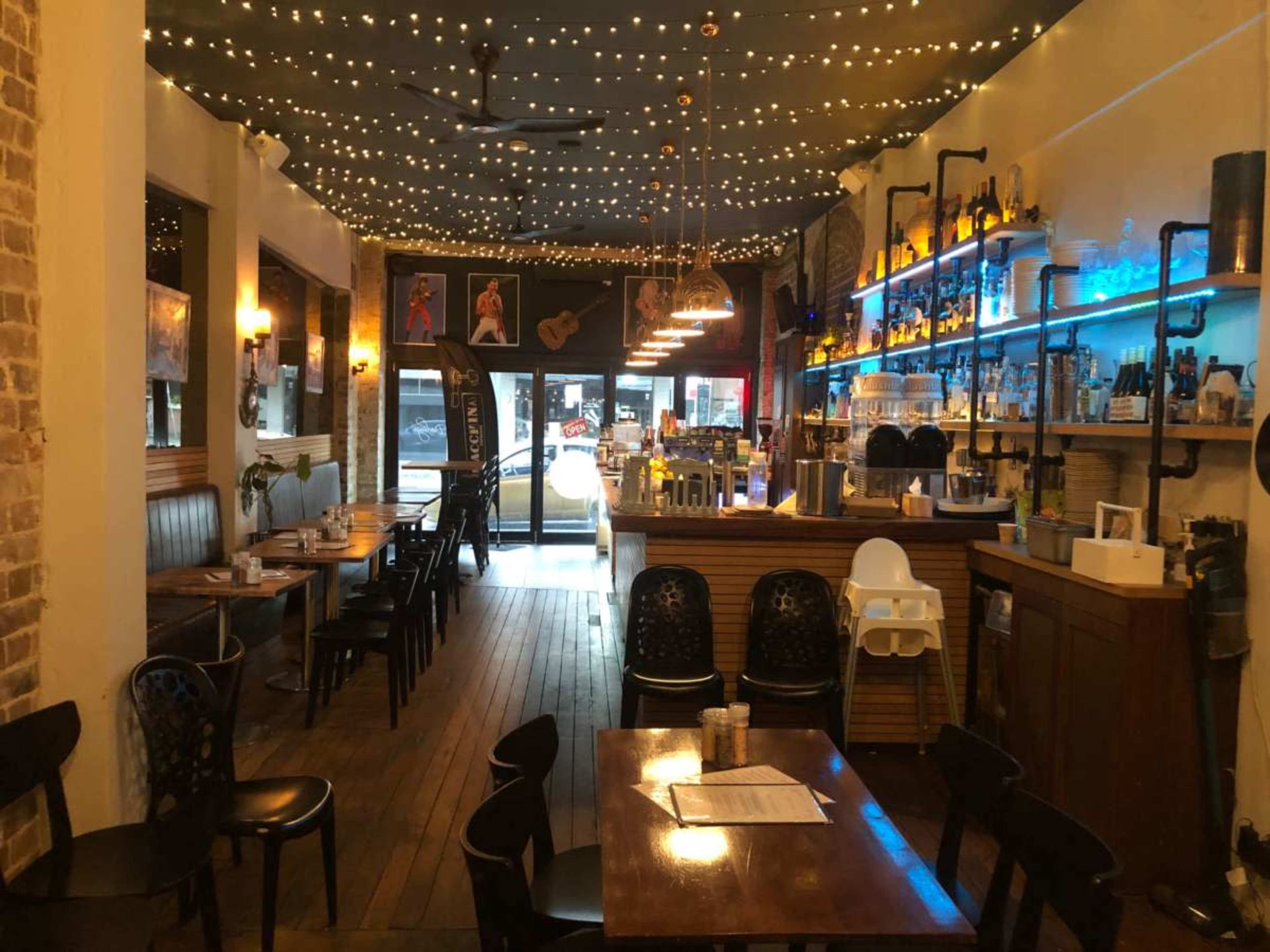 The image shows a restaurant interior with exposed brick walls, wooden floors, a bar with colorful backlighting, and various seating arrangements under twinkling overhead lights.