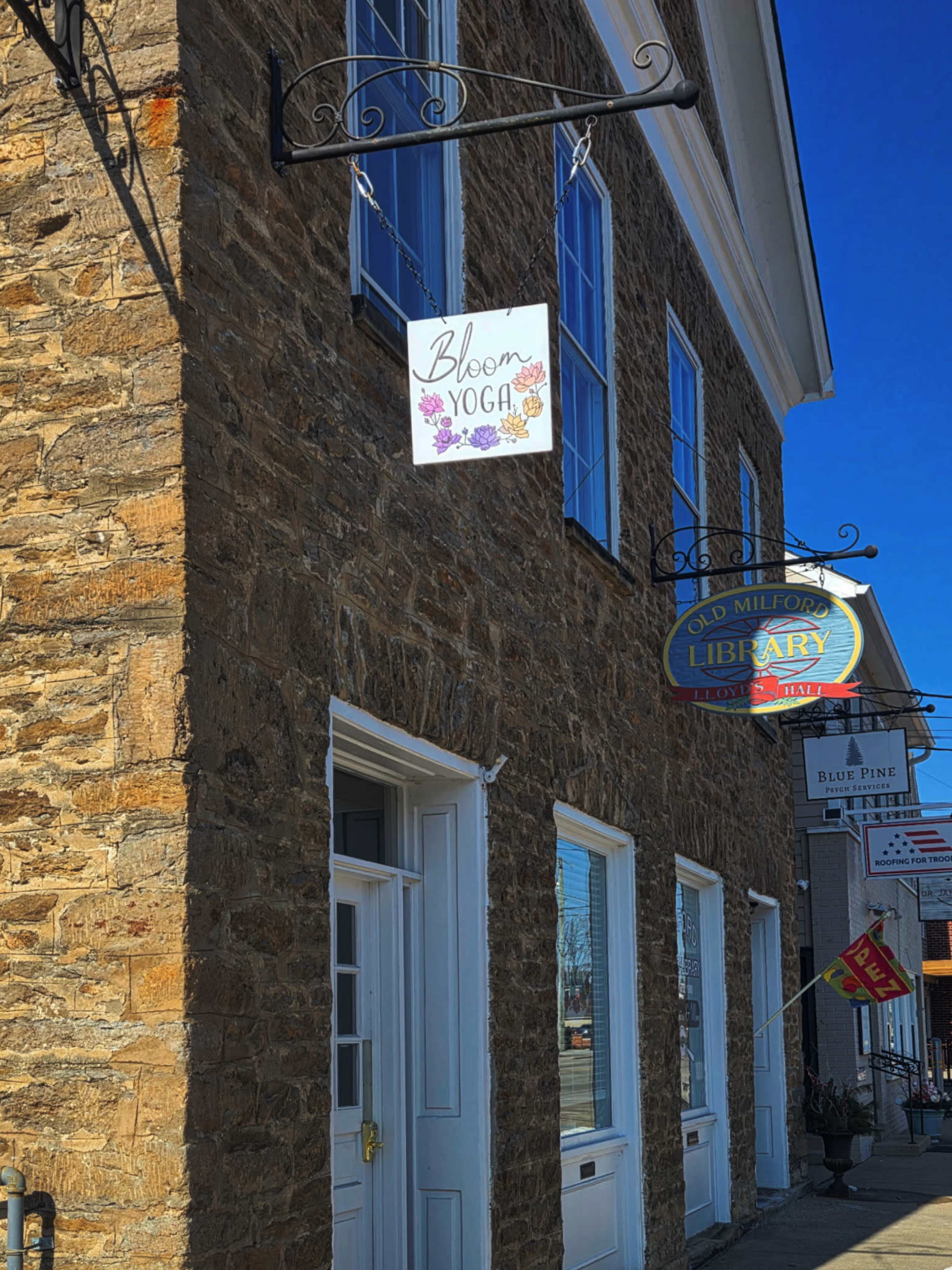 The image shows a stone building with two signs, one for Bloom Yoga and the other for the Old Milford Library.