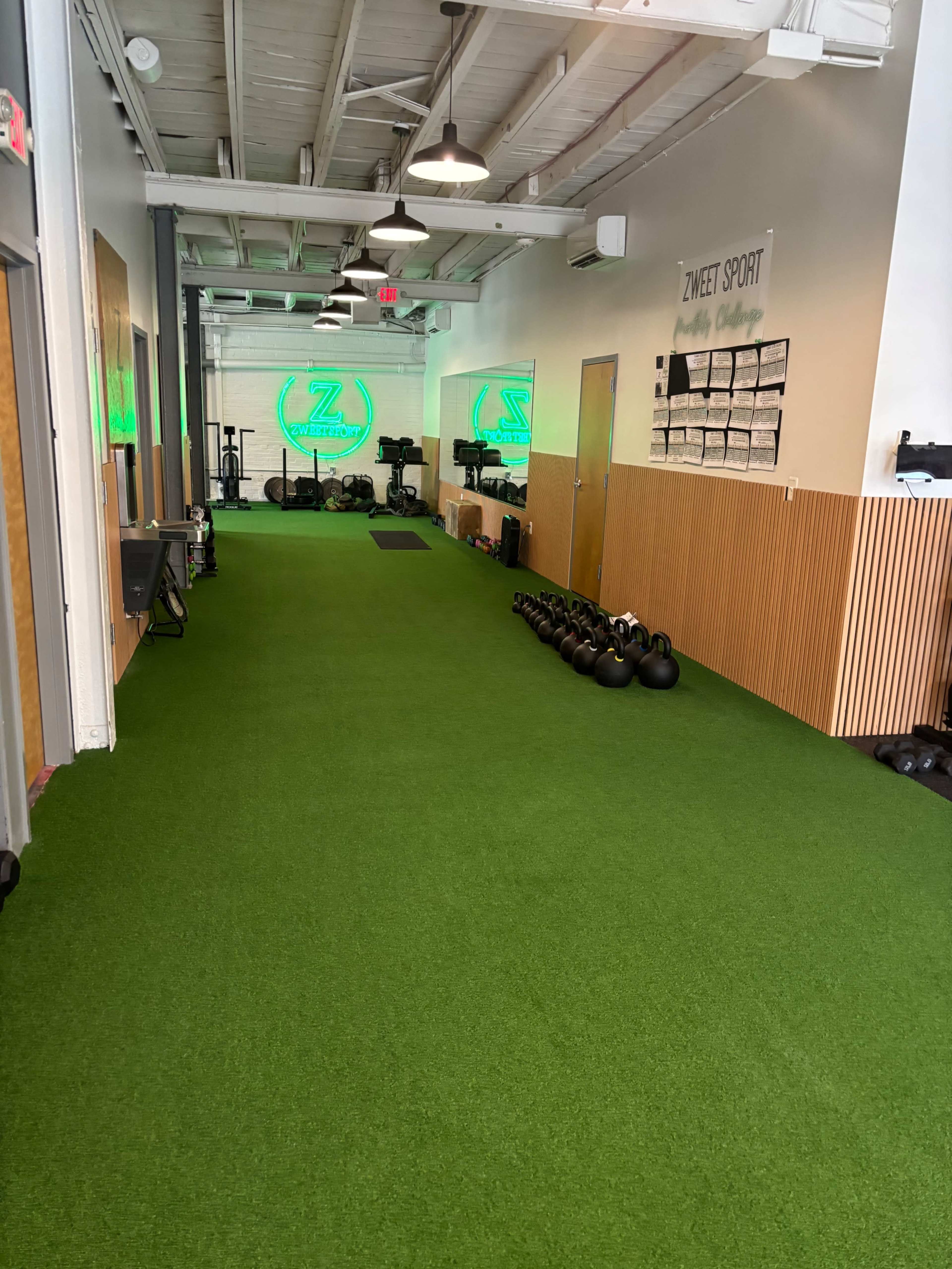 The image shows a fitness studio with a long, green artificial turf area, kettlebells lined against the wall, and workout equipment in the background.
