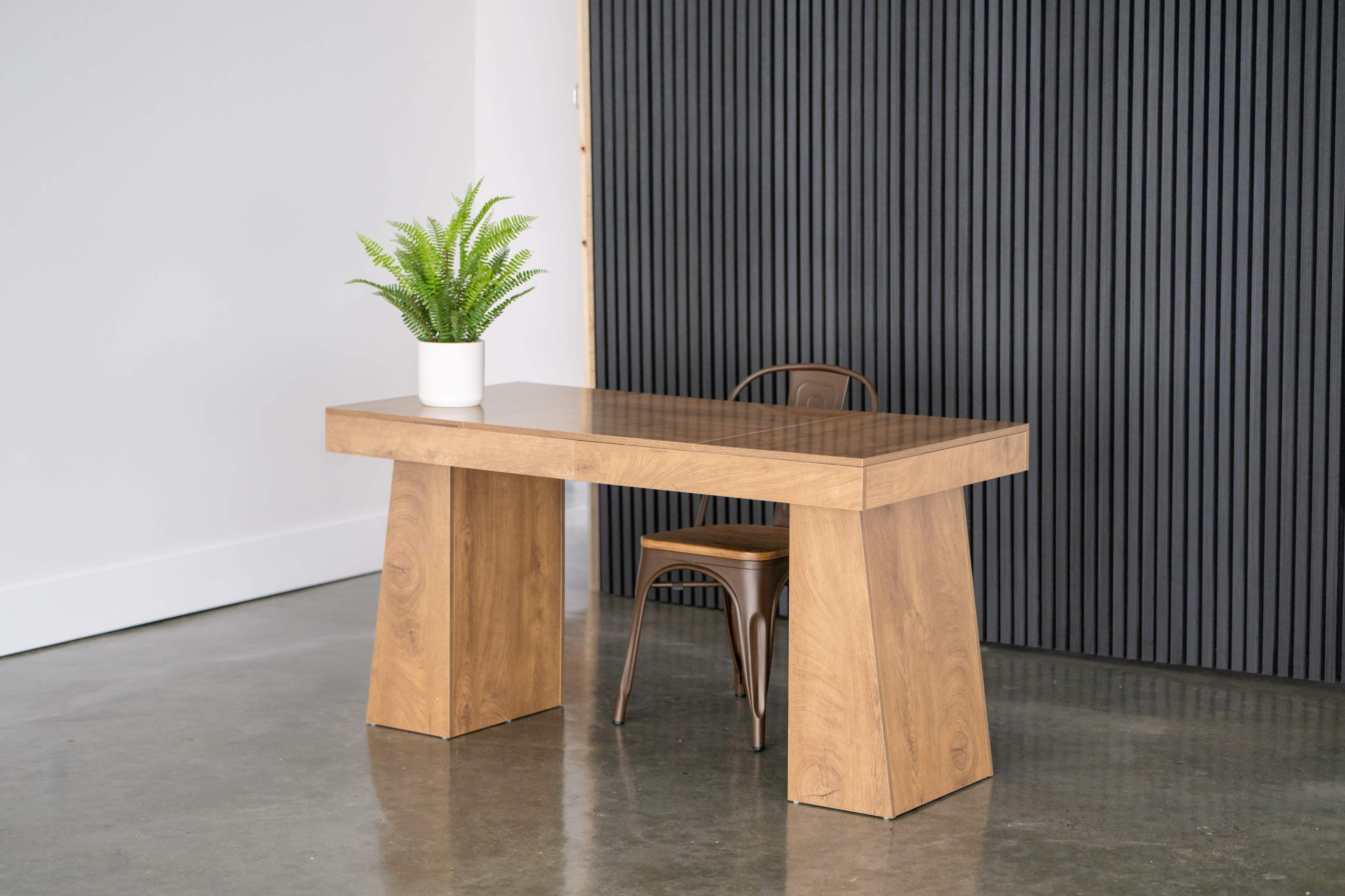 A wooden table with a minimalist design is set against a black textured wall, accompanied by a single chair and a potted plant.