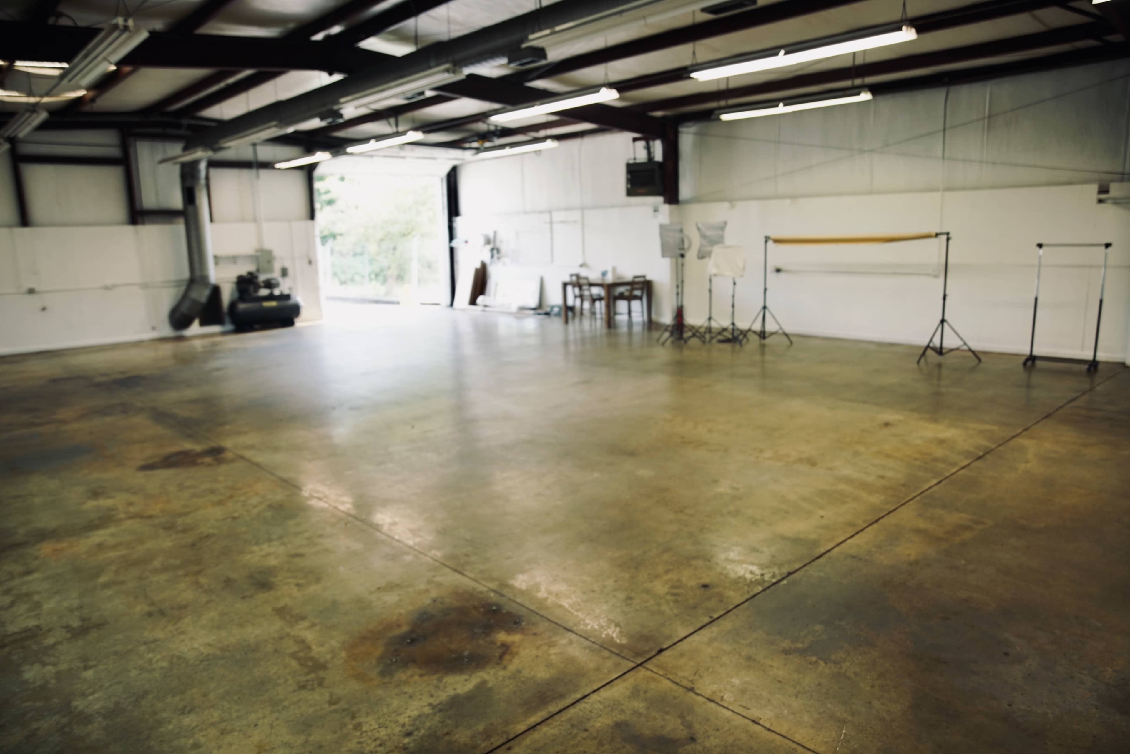 The image shows a spacious, empty warehouse with concrete floors and sparse equipment, including a table and photography stands, set against a backdrop of white walls.