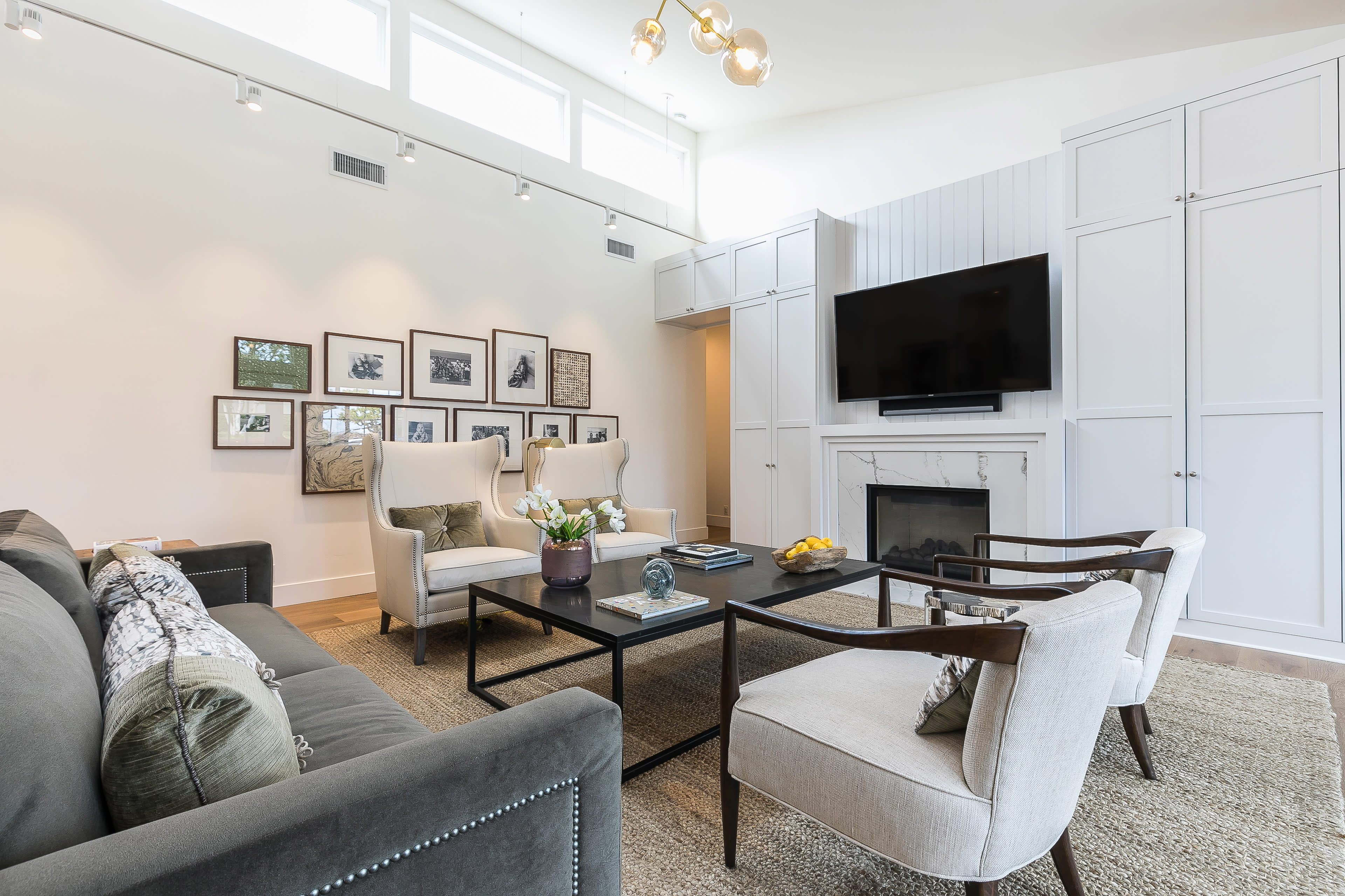 A modern living room features a sofa, two armchairs, a coffee table, and a wall-mounted television, with framed photos arranged above a console table.