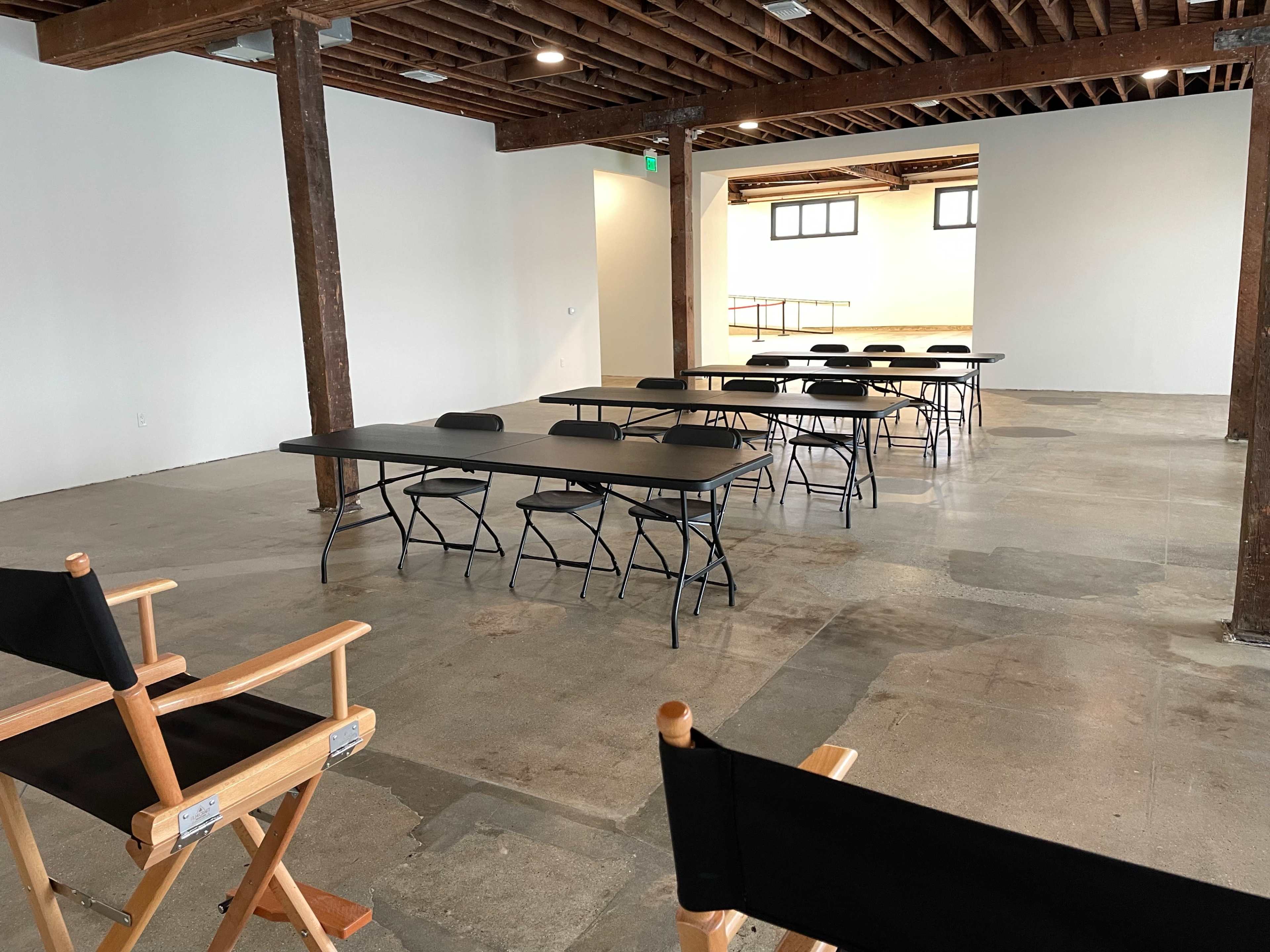 An open, empty room features multiple rows of black tables and chairs, with director's chairs positioned in the foreground.