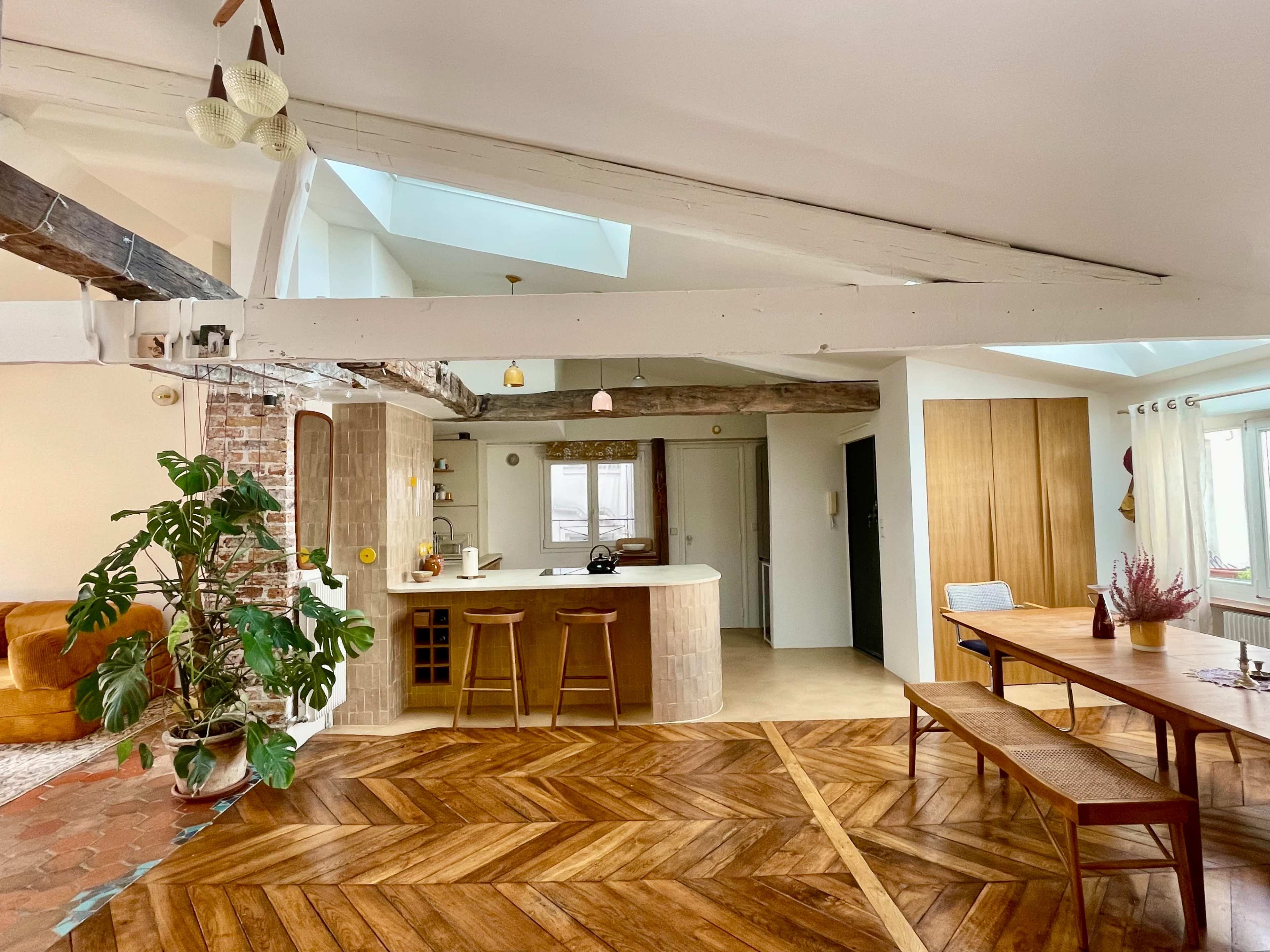 The image shows a spacious, open-concept living area with a kitchen island, dining table, and wood-paneled flooring, complemented by large windows and natural light.
