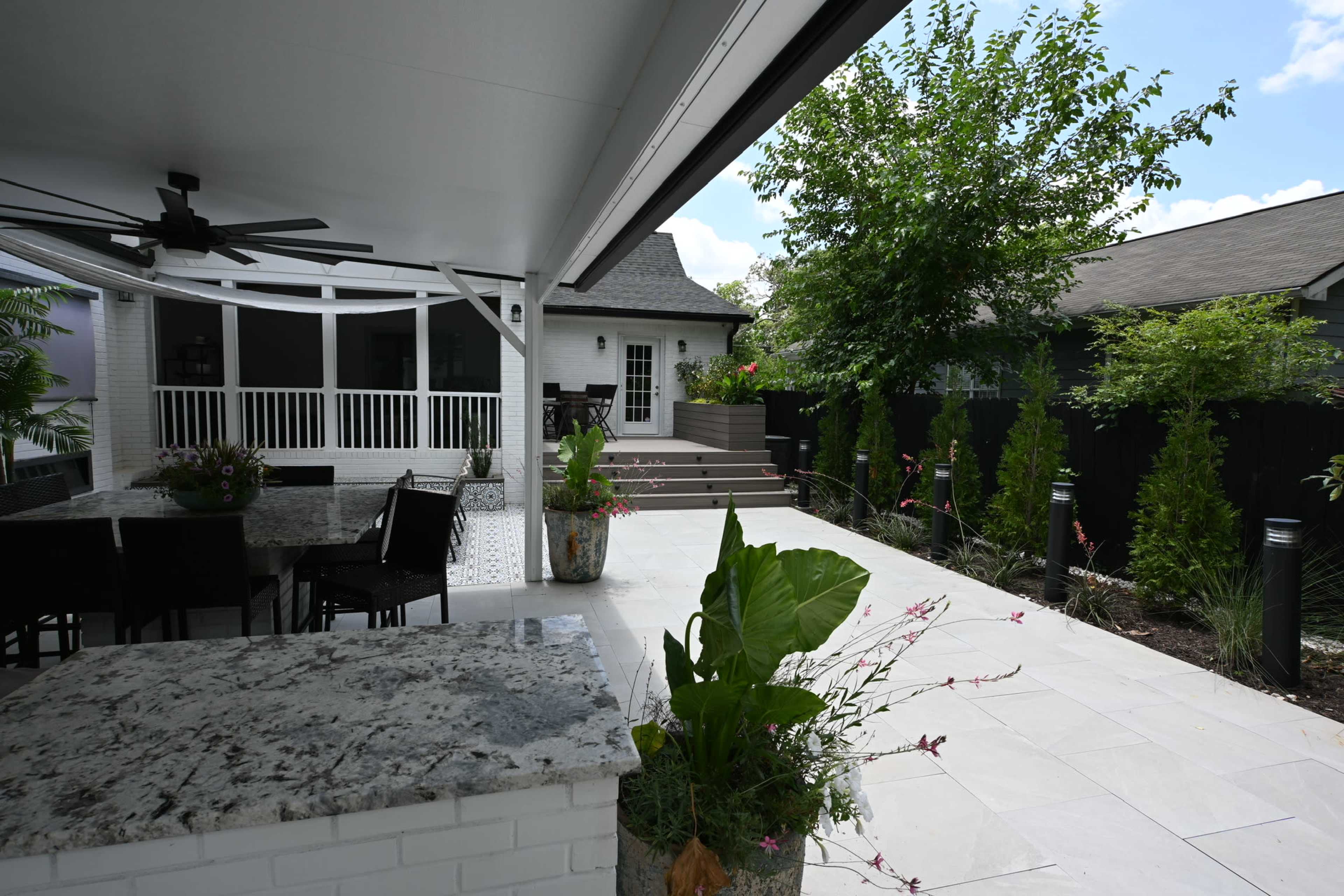 The image shows a spacious outdoor patio with a granite countertop, dining area, and surrounding greenery, including trees and flower beds.